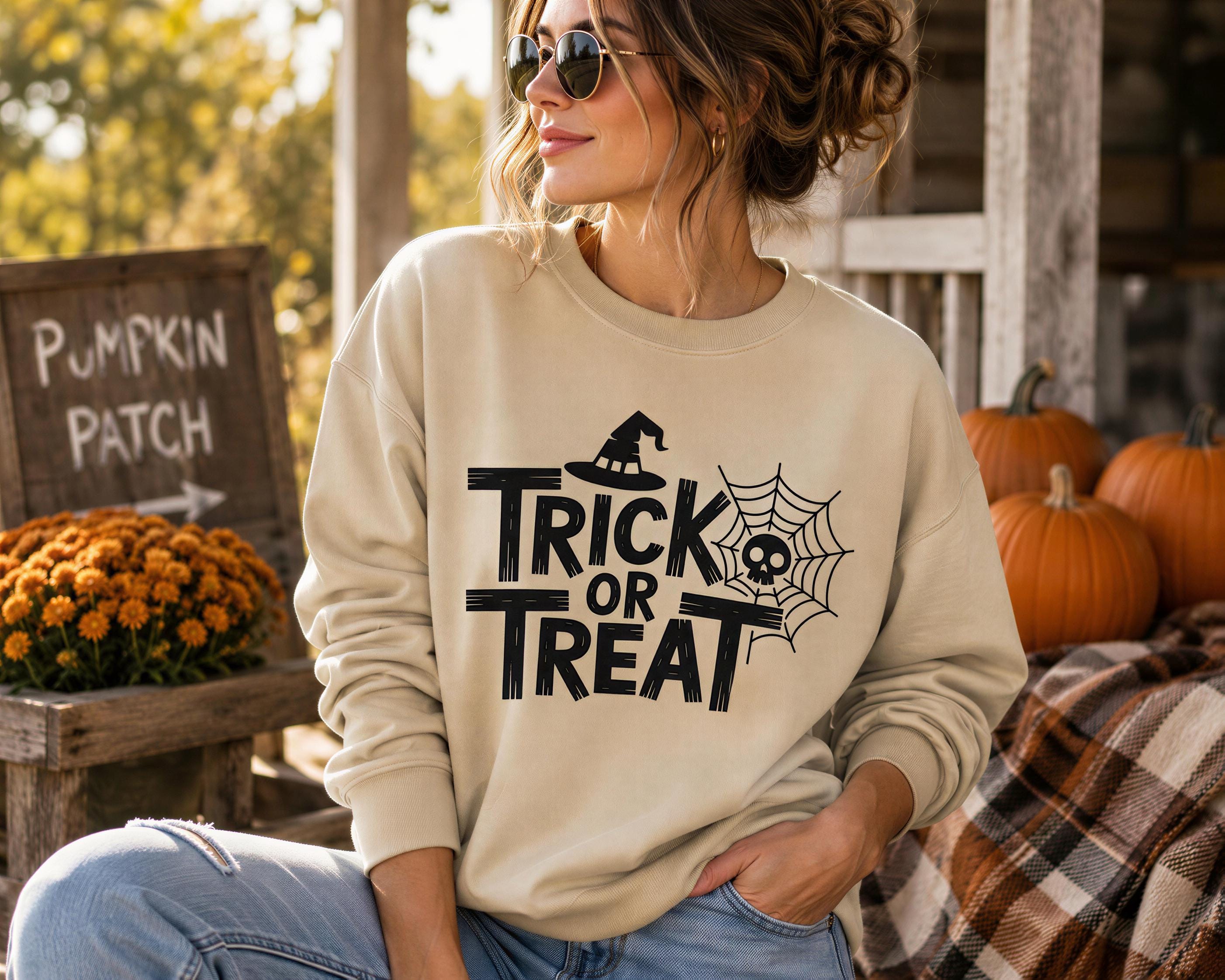 A woman wearing a beige sweater with the text "TRICK OR TREAT" and sunglasses is sitting in front of a pumpkin patch with pumpkins and flowers.