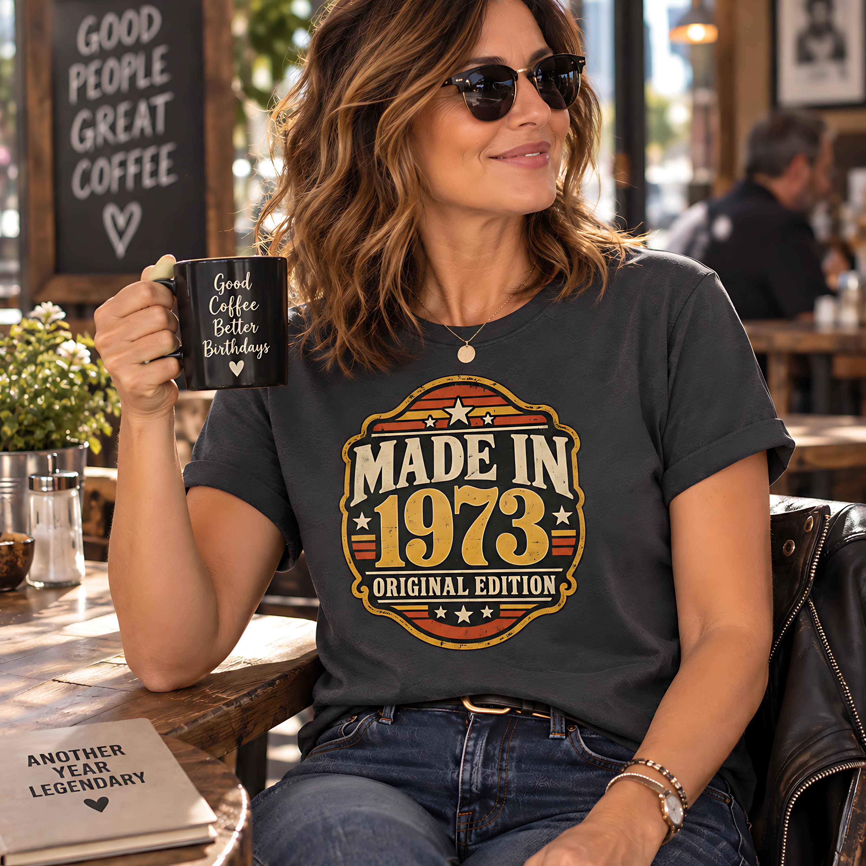 A woman wearing sunglasses and a t-shirt with a graphic design is sitting at a table in a cafe, holding a coffee mug and smiling.