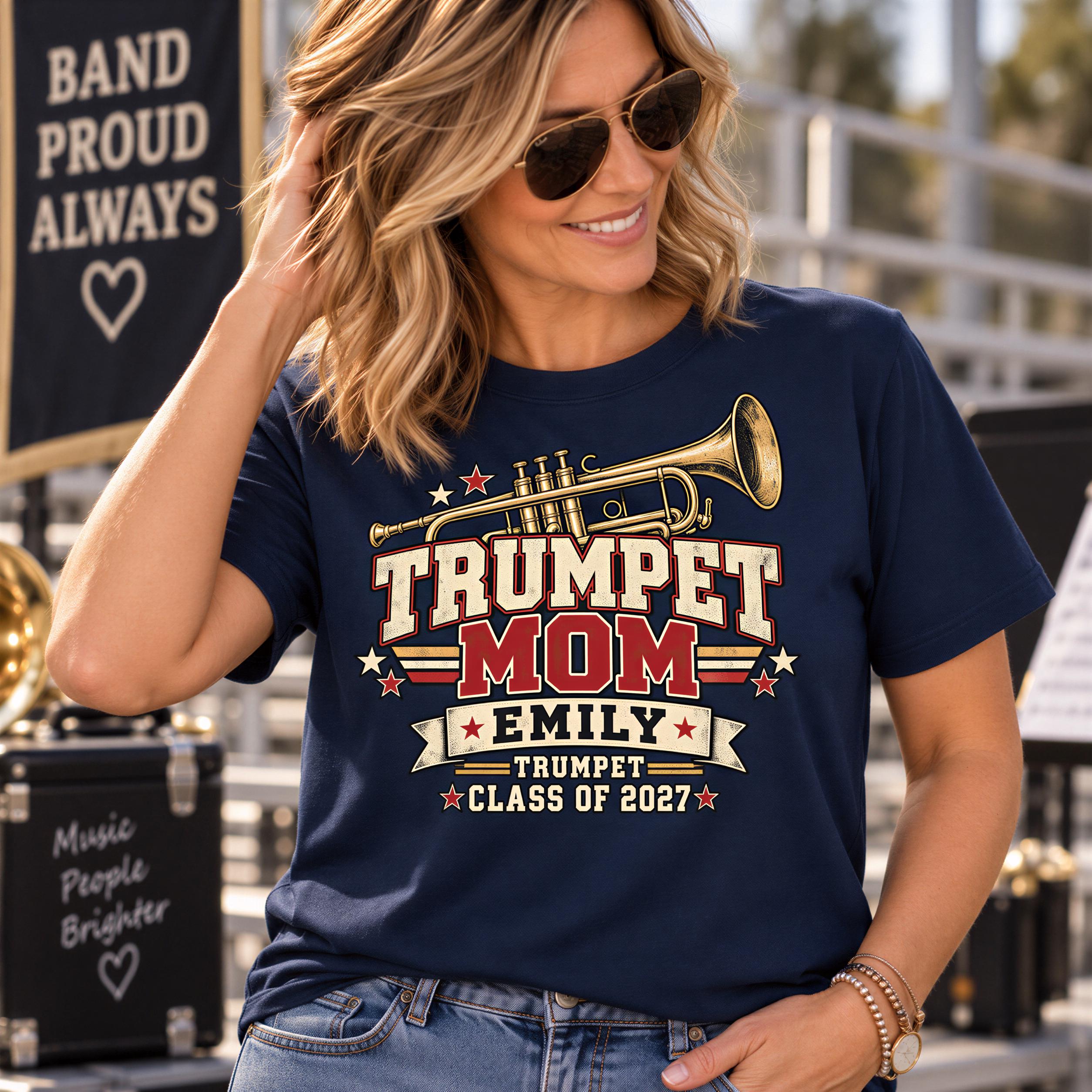 A woman wearing sunglasses and a navy blue t-shirt with a trumpet graphic and text, smiling and posing in front of a banner that says "BAND PROUD ALWAYS".