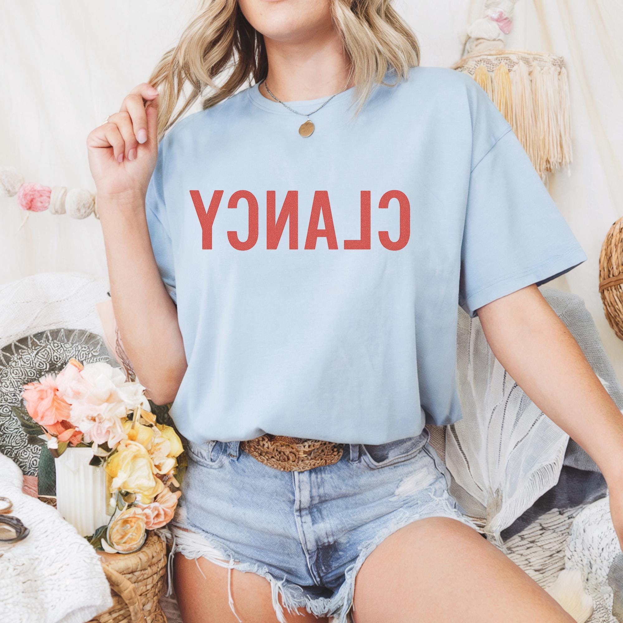 A woman wearing a light blue t-shirt with the word "CLAN" printed on it, paired with denim shorts, sits in a casual setting with a basket of flowers and a woven basket nearby.