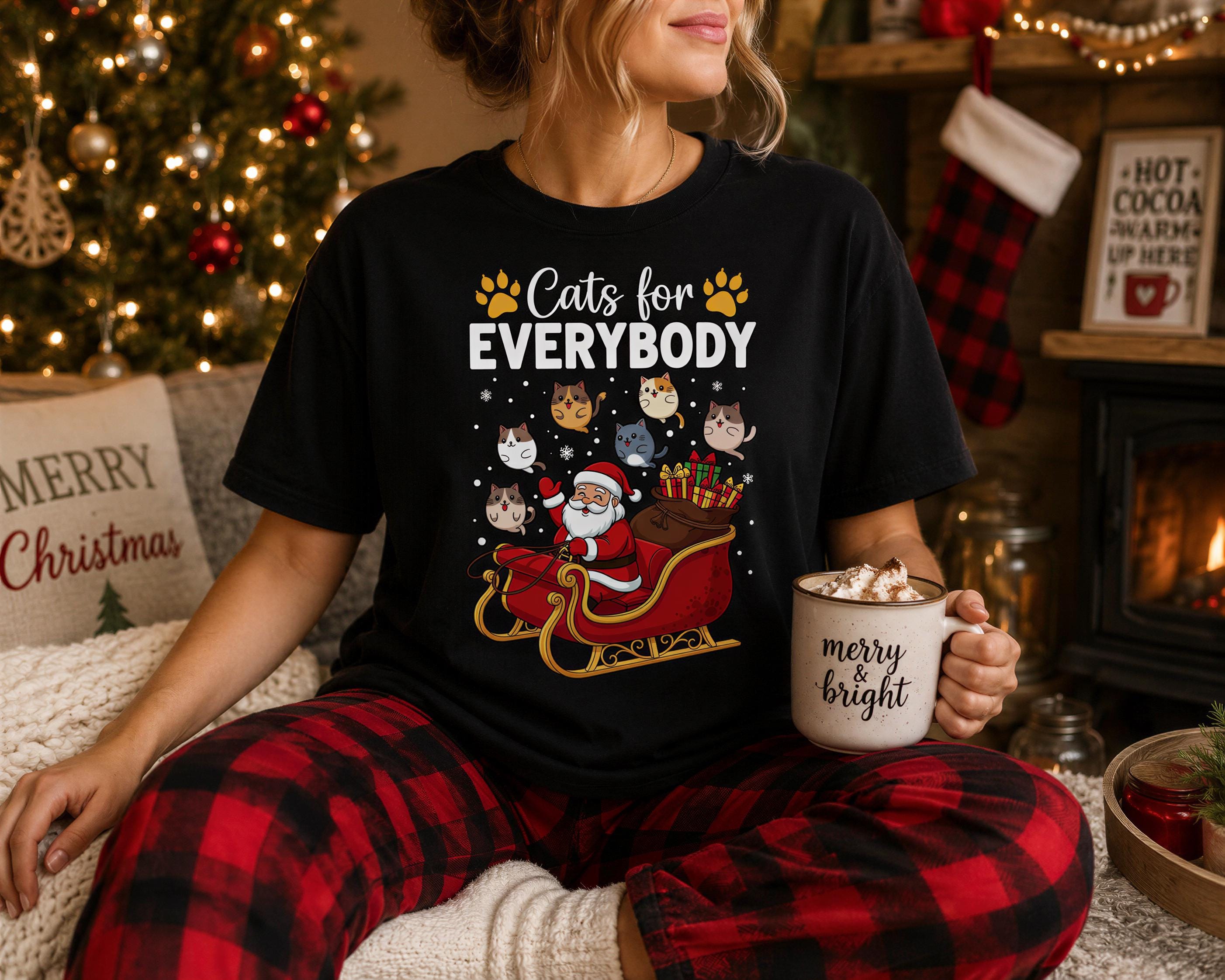 A woman wearing a black t-shirt with a graphic of Santa Claus and cats on a sled, holding a mug with the words "Merry & Bright" written on it, sitting in front of a Christmas tree and a fireplace.
