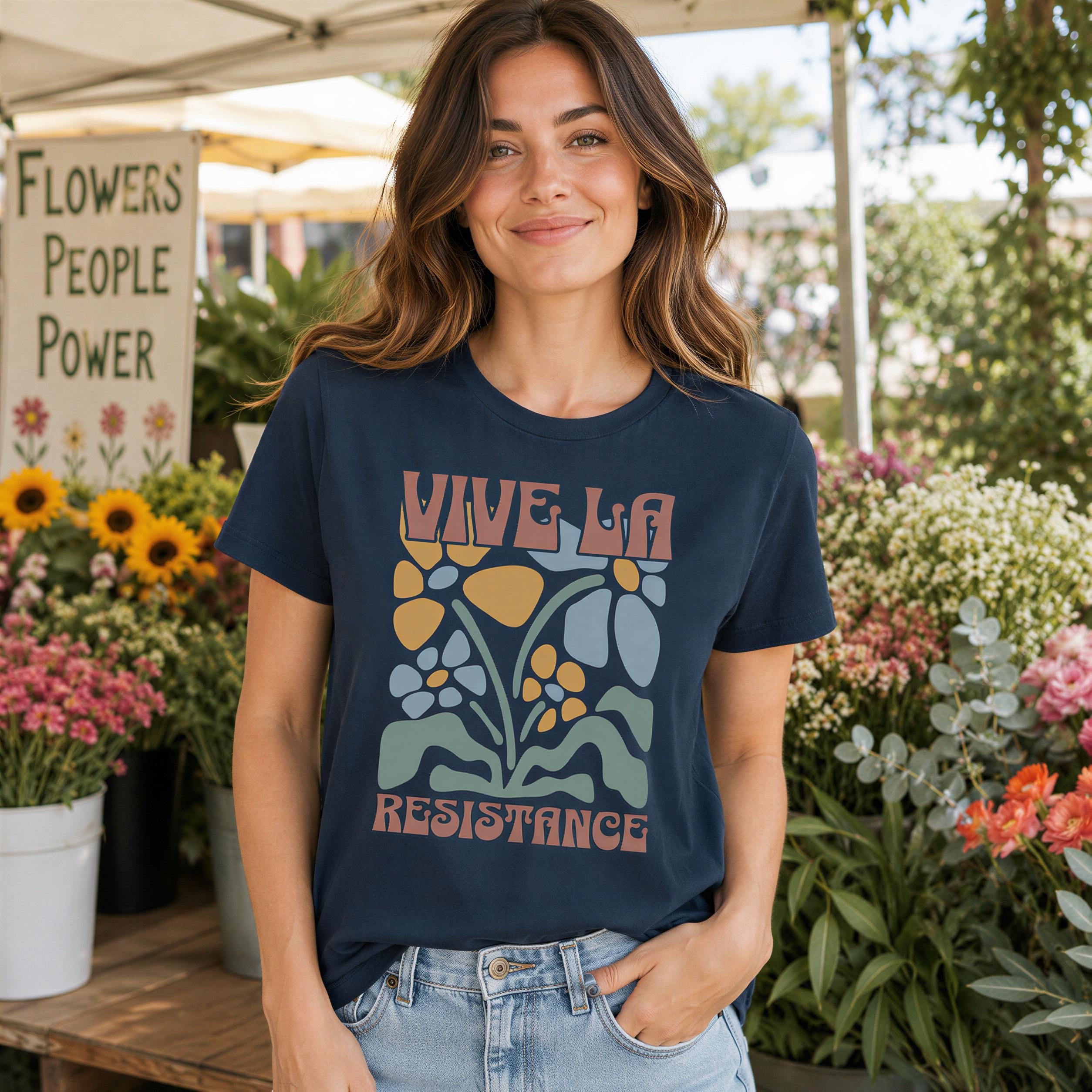 A woman wearing a blue t-shirt with a graphic design stands in front of a flower stand, smiling at the camera.