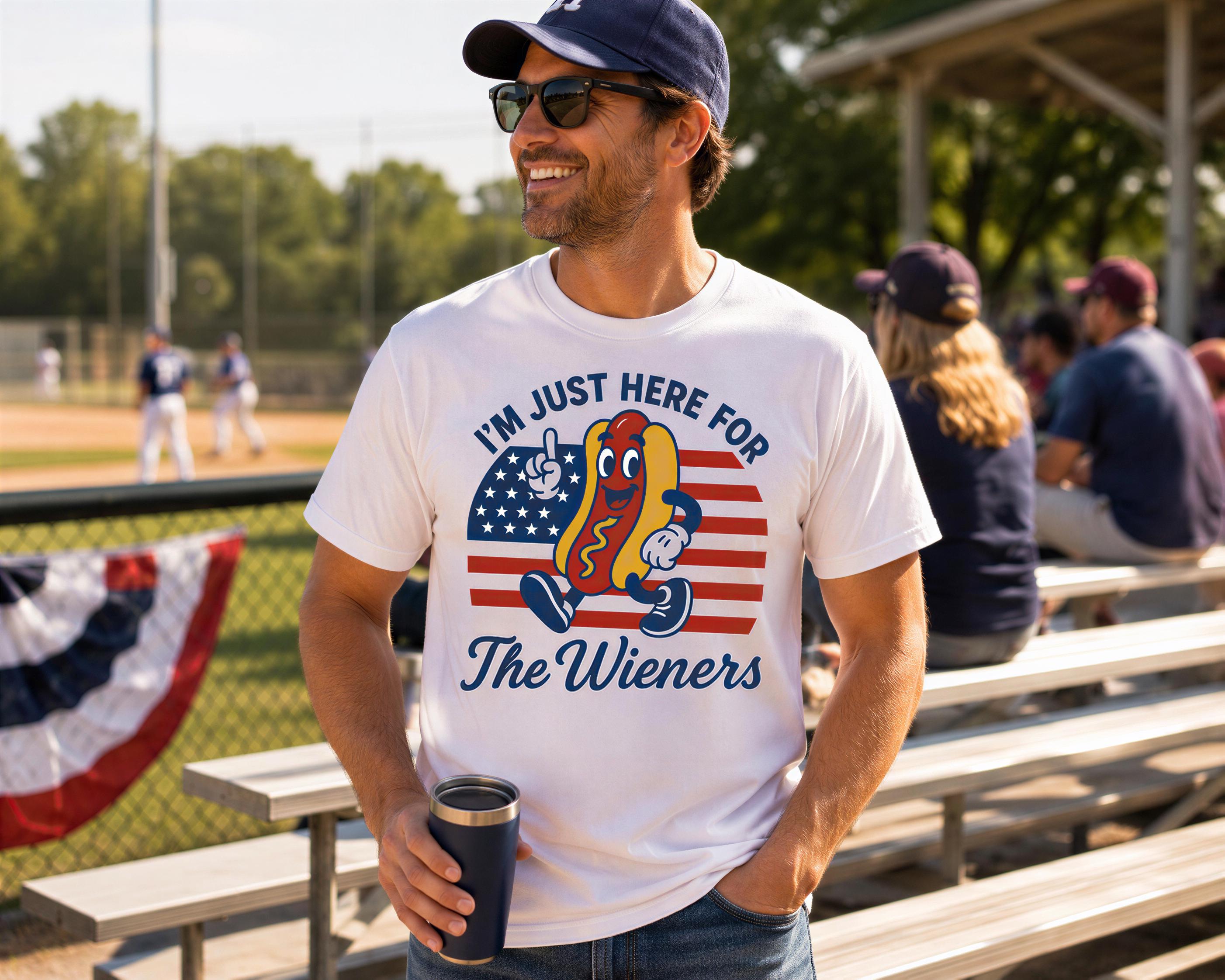 A man wearing a white t-shirt with a graphic of a hot dog and the text "I'M JUST HERE FOR The Wieners" is standing in a baseball stadium, holding a cup.