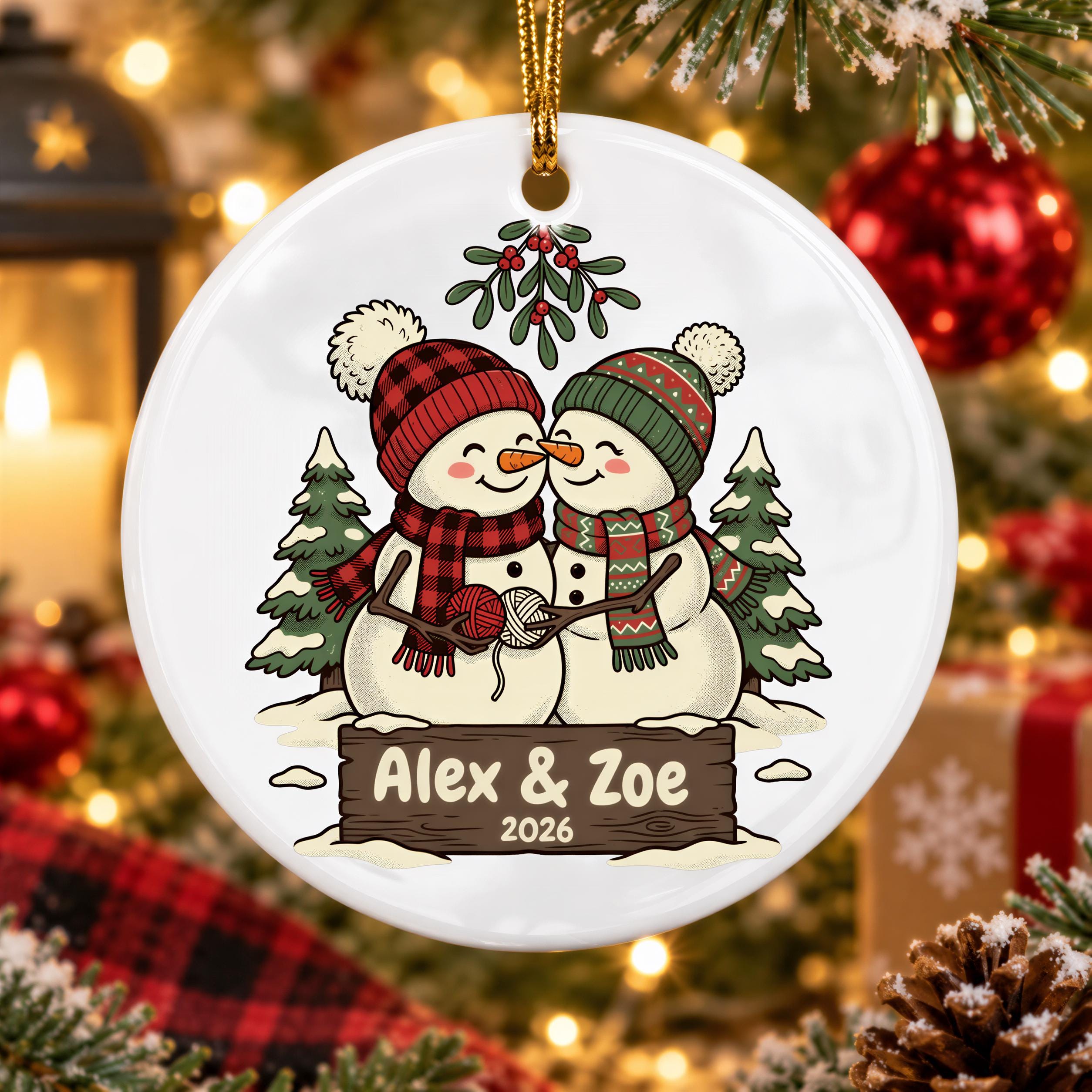 A white ornament with a snowman and two snowmen, along with a sign that says "Alex & Zoe" and the year "2026". The ornament is hanging on a Christmas tree, surrounded by other holiday decorations.