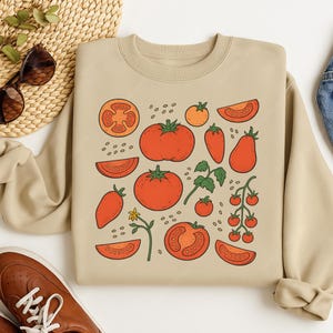 May include: Beige sweatshirt featuring a repeating pattern of red tomatoes, tomato slices, and small orange tomatoes. The design includes green leaves and small yellow flowers. The tomatoes are outlined in black.