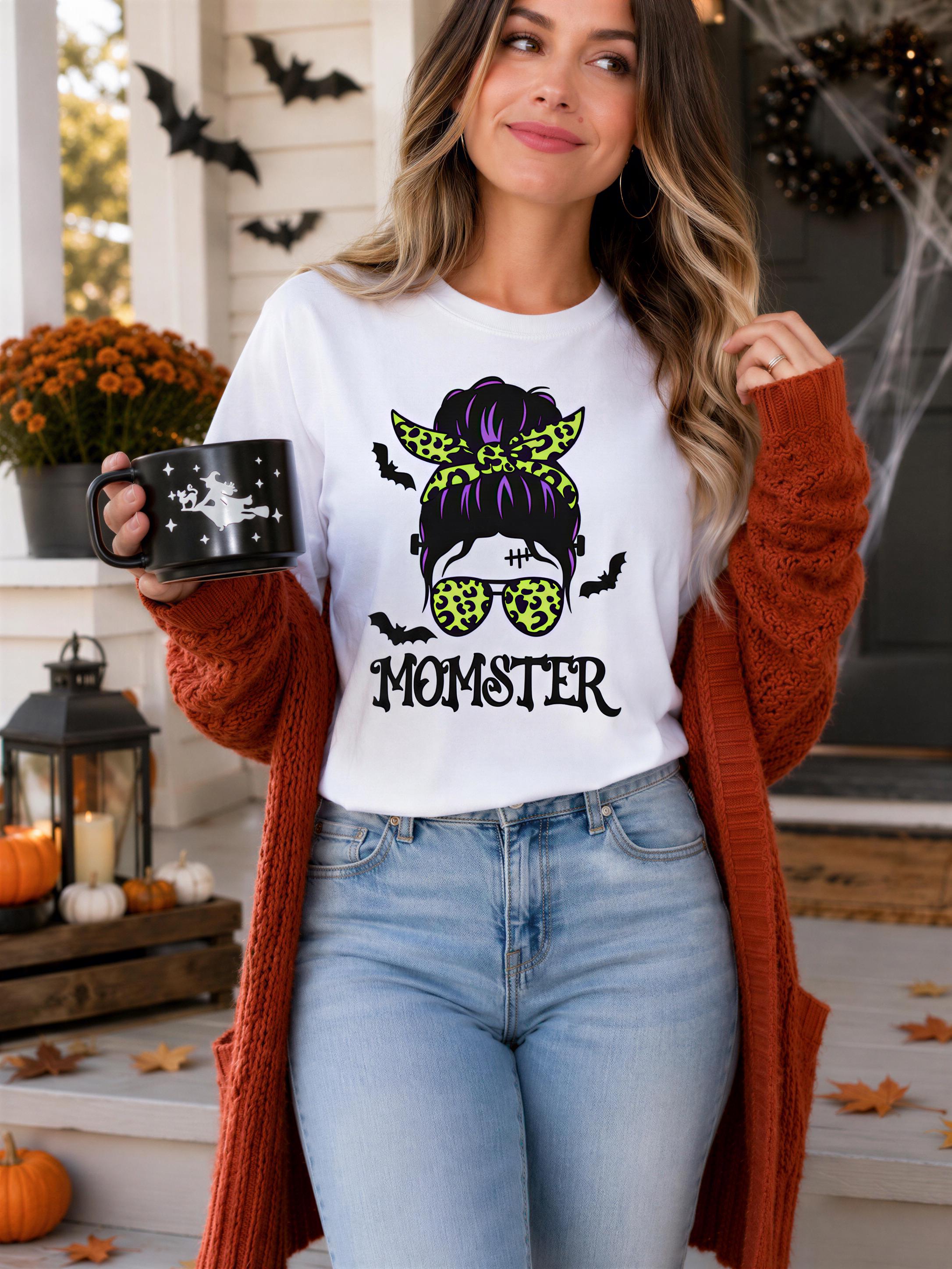 A woman wearing a white t-shirt with a graphic of a woman's face and the word "MOMSTER" printed on it, holding a black mug with a star design. She is standing on a porch decorated for Halloween, with a lantern, pumpkins, and bats visible in the background.