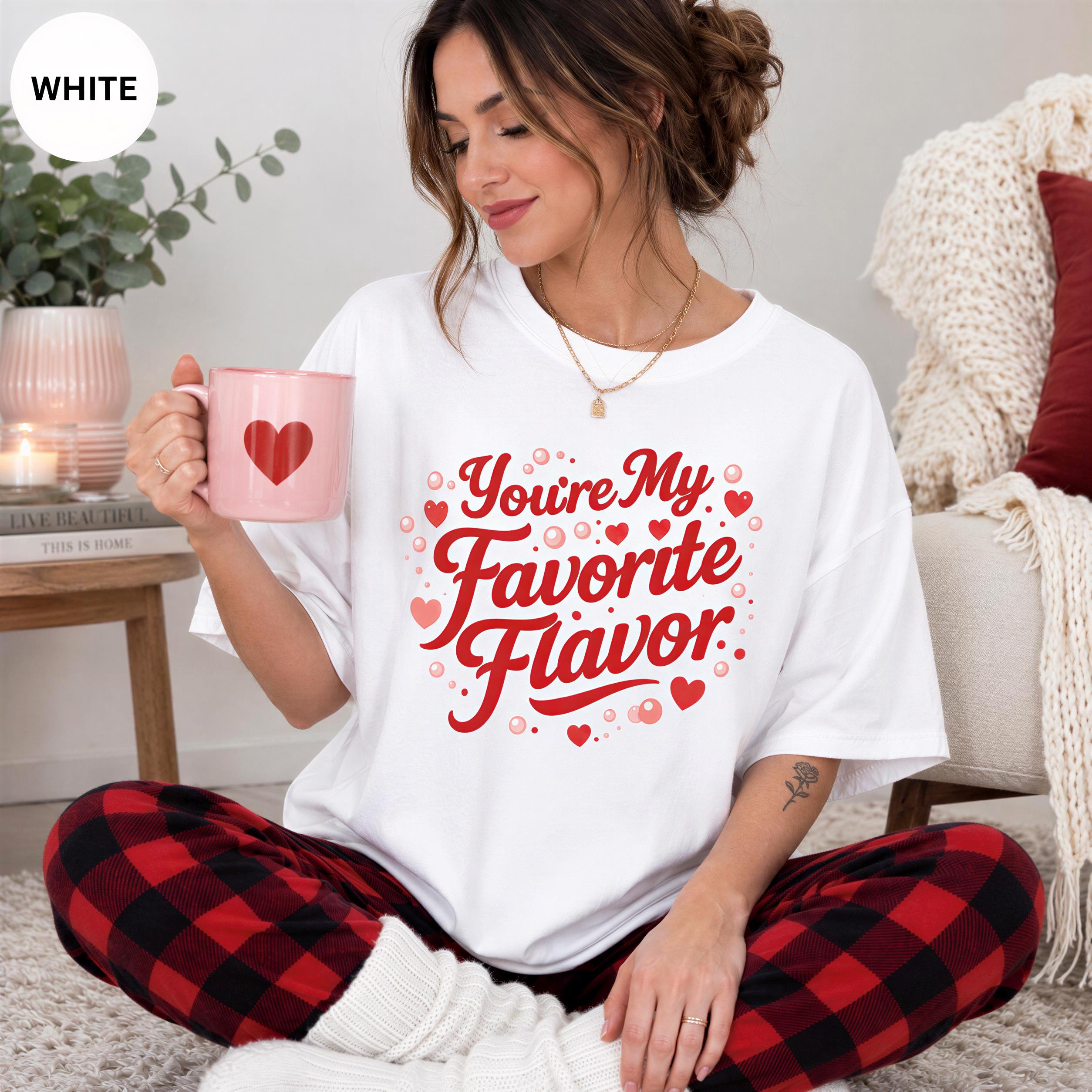 A woman wearing a white t-shirt with a red heart design and the text "You're My Favorite Flavor" is sitting on the floor, holding a pink mug.