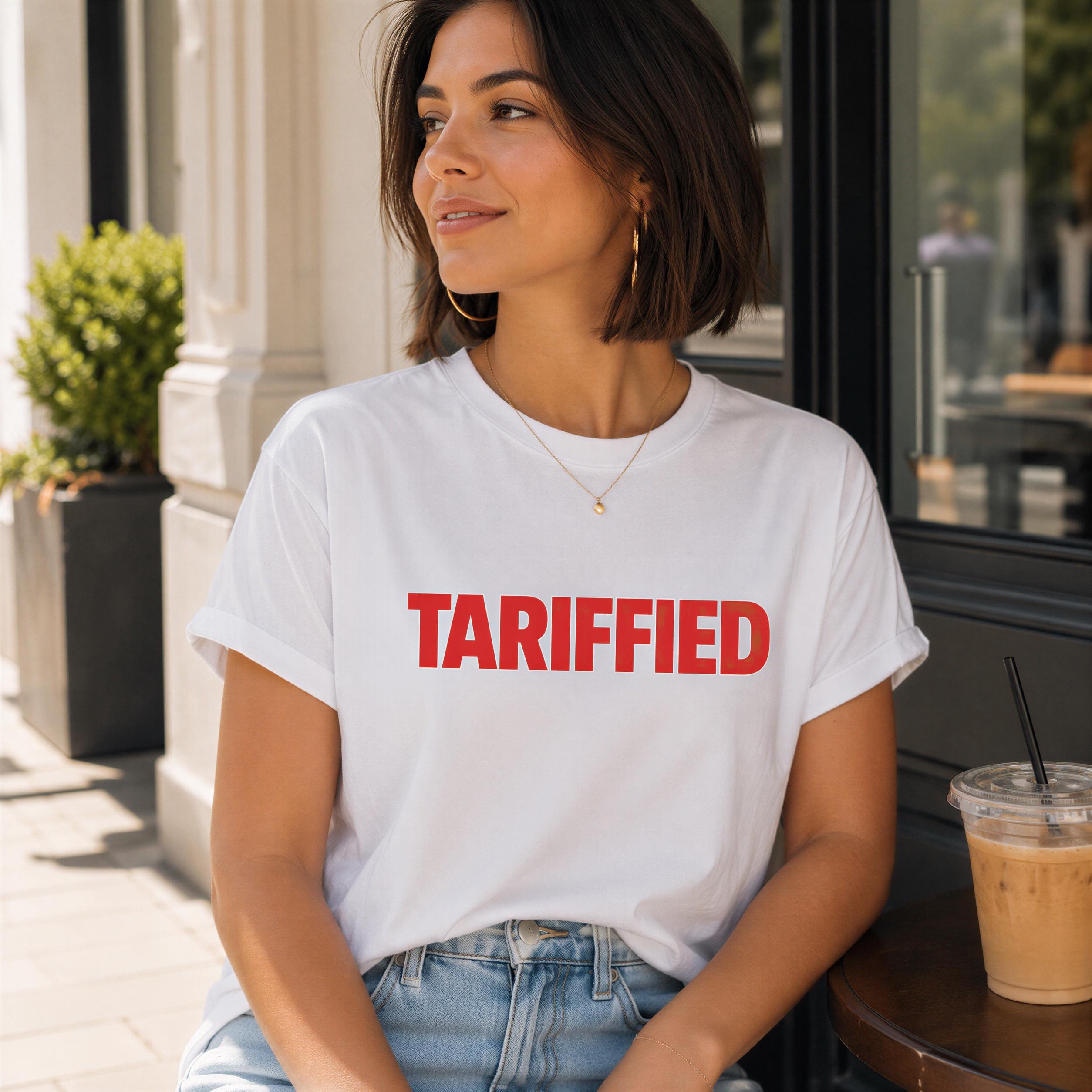A woman wearing a white t-shirt with the word "TARIFFED" printed on it, sitting at a table with a drink in a plastic cup.