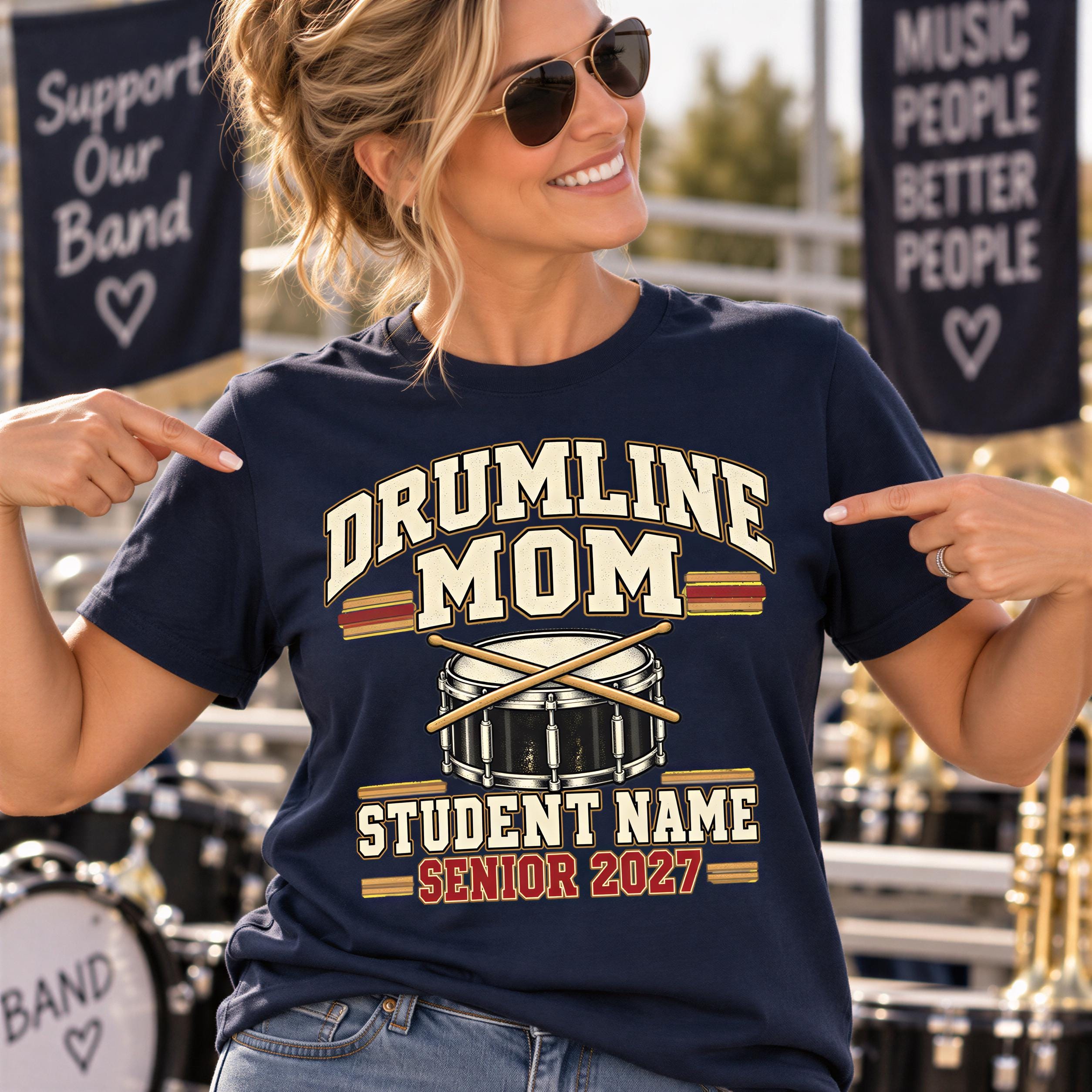 A smiling woman wearing a navy blue t-shirt with a drumline design and text, standing in front of a banner that says "Support Our Band" and "Music People Better People".
