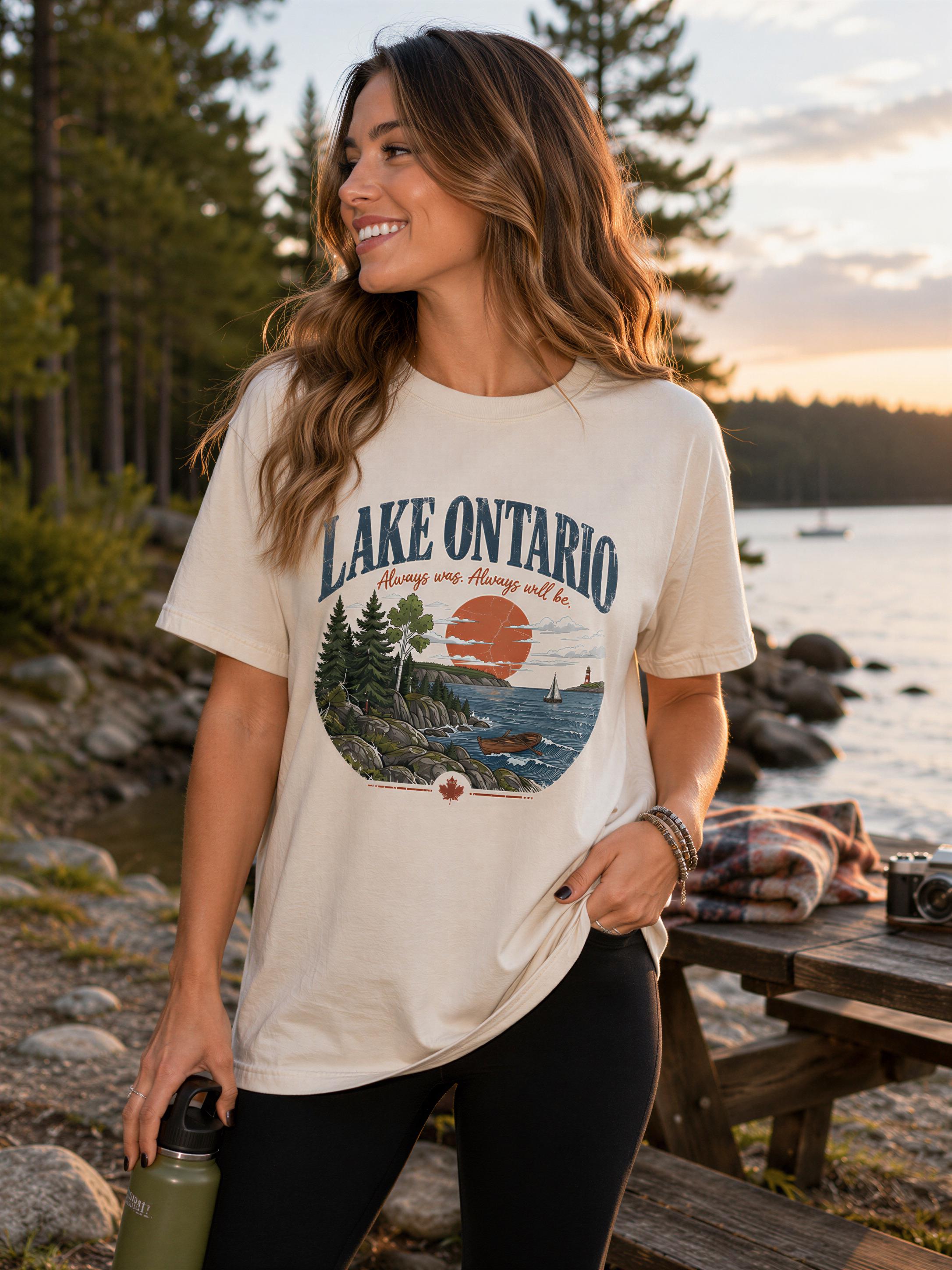 A woman wearing a t-shirt with a lake Ontario graphic stands near a picnic table, smiling and holding a water bottle.