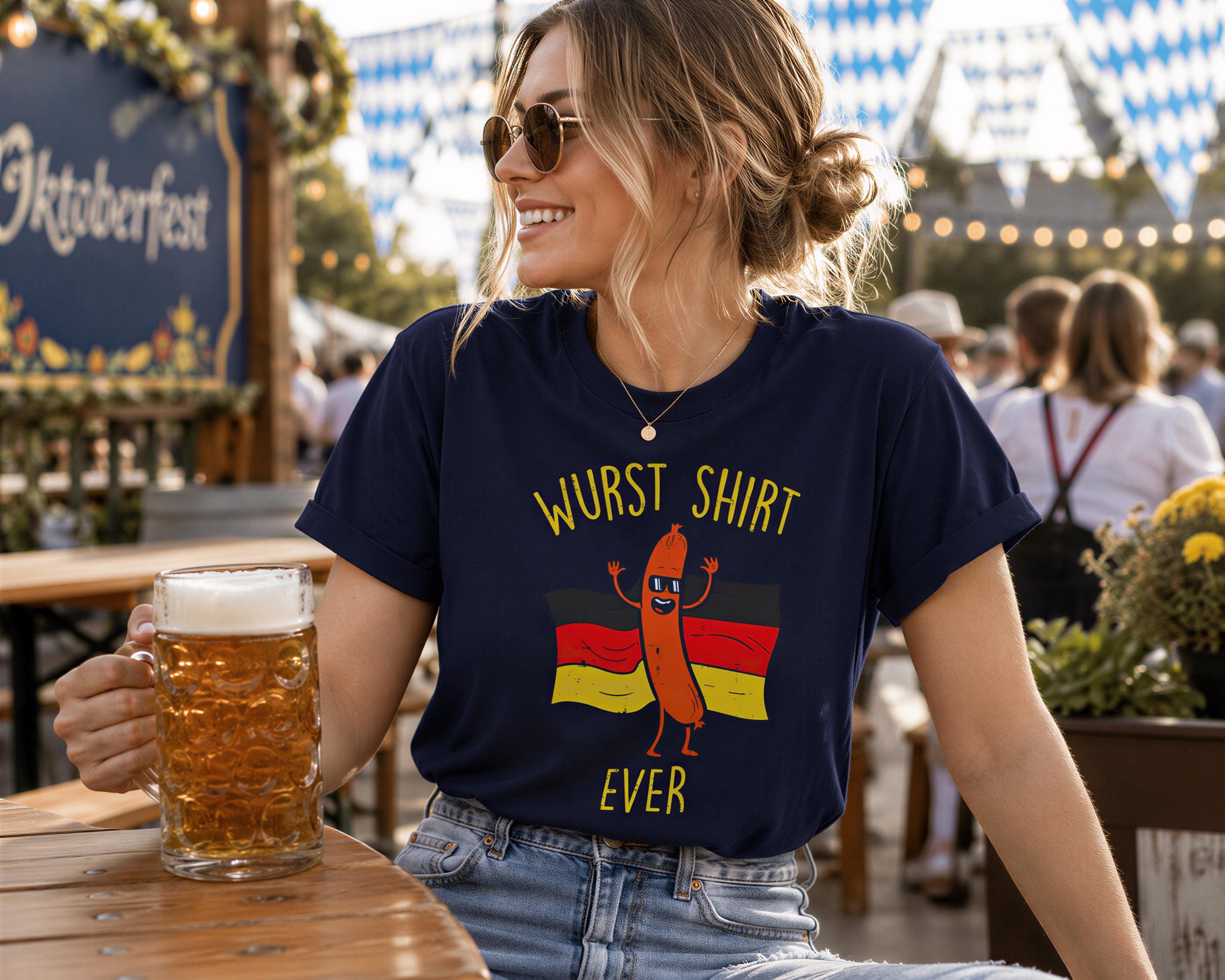 A woman wearing a blue t-shirt with a cartoon hot dog and the text "Würst Shirt Ever" is sitting at a table with a glass of beer in front of her.