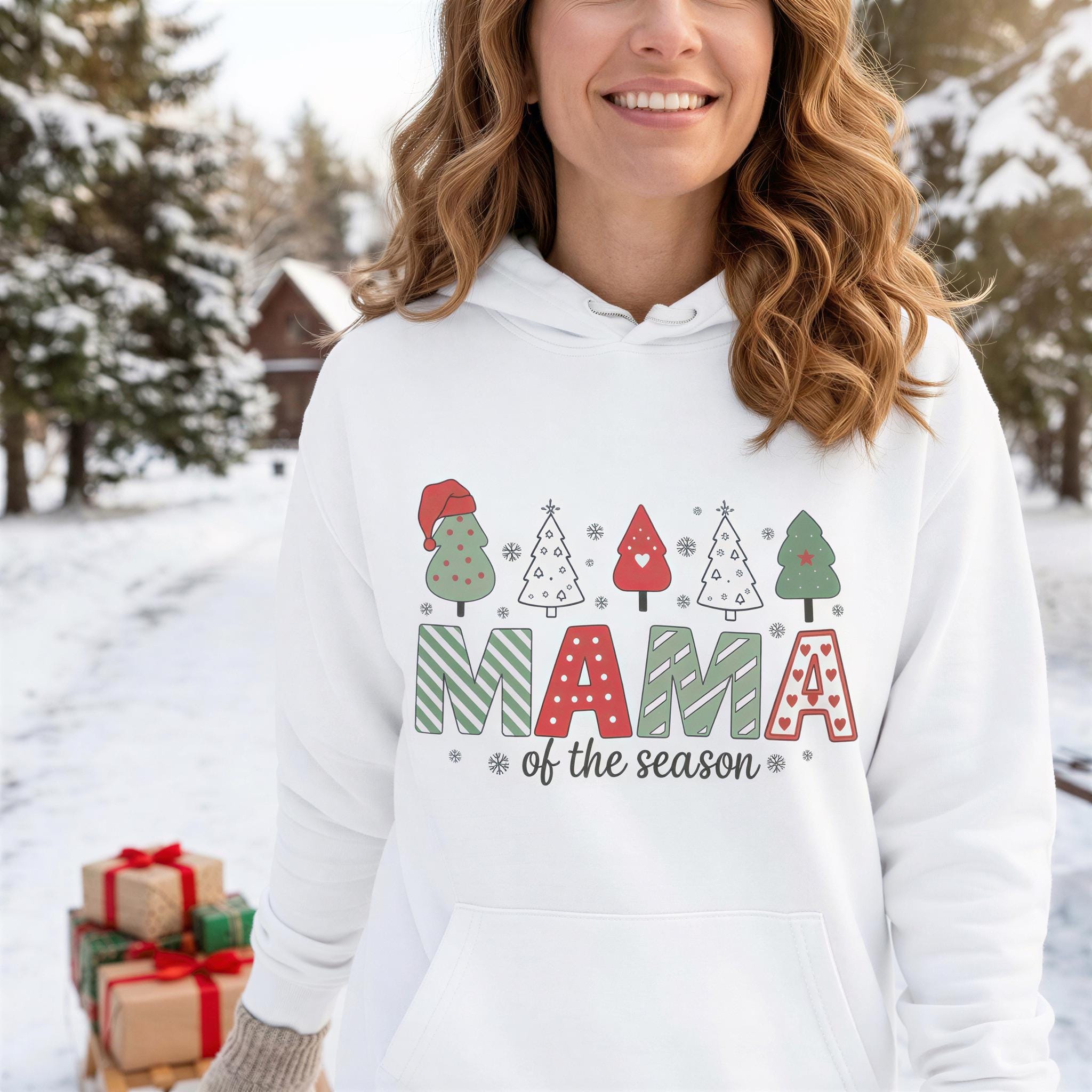 A woman wearing a white hoodie with a graphic design of Christmas trees and the text "MAMA of the season" printed on it, smiling in a snowy outdoor setting.