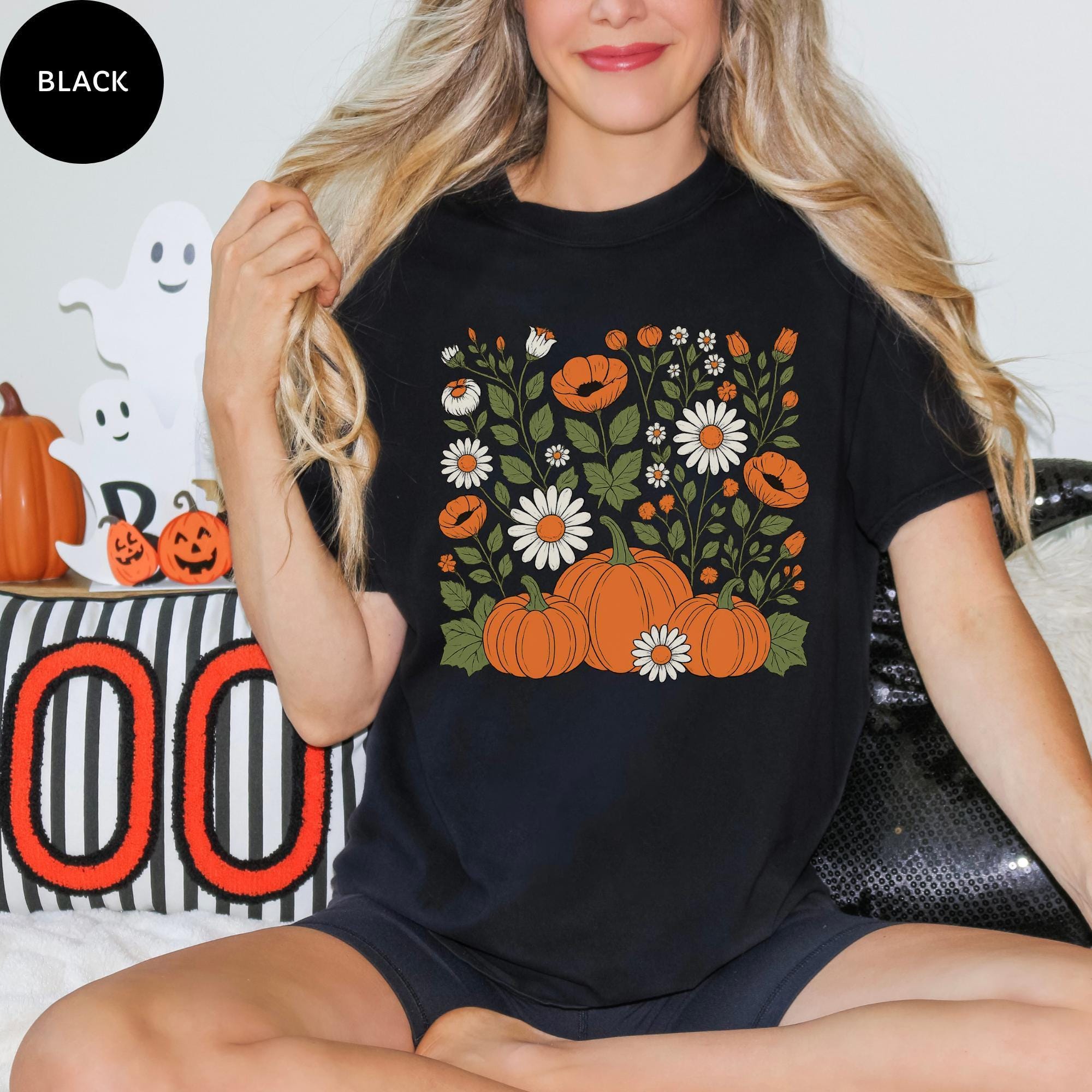 A woman wearing a black t-shirt with a graphic design of pumpkins and flowers, sitting on a couch with Halloween decorations in the background.