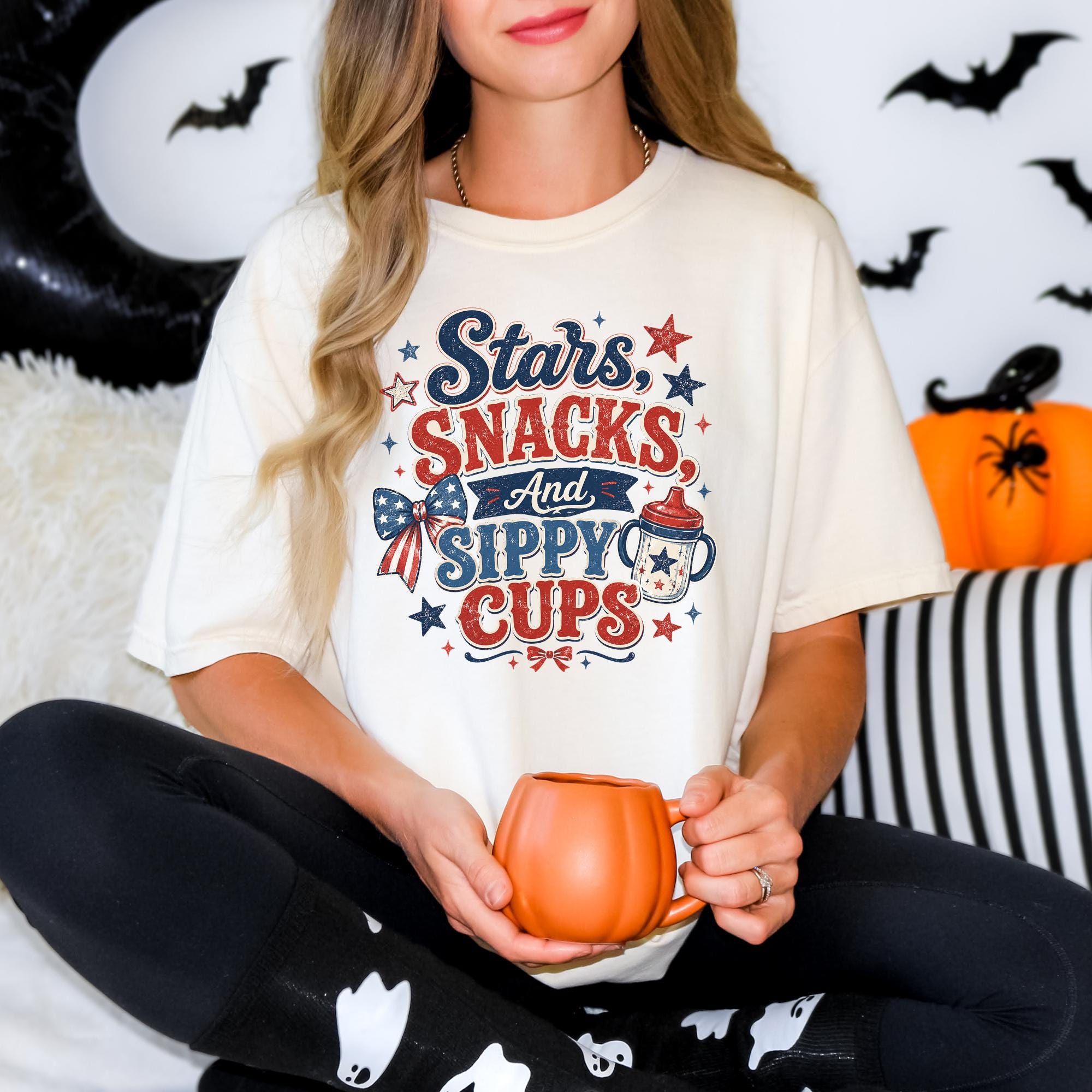 A woman wearing a white t-shirt with a graphic design and holding a pumpkin-shaped mug, sitting on a couch with Halloween decorations in the background.