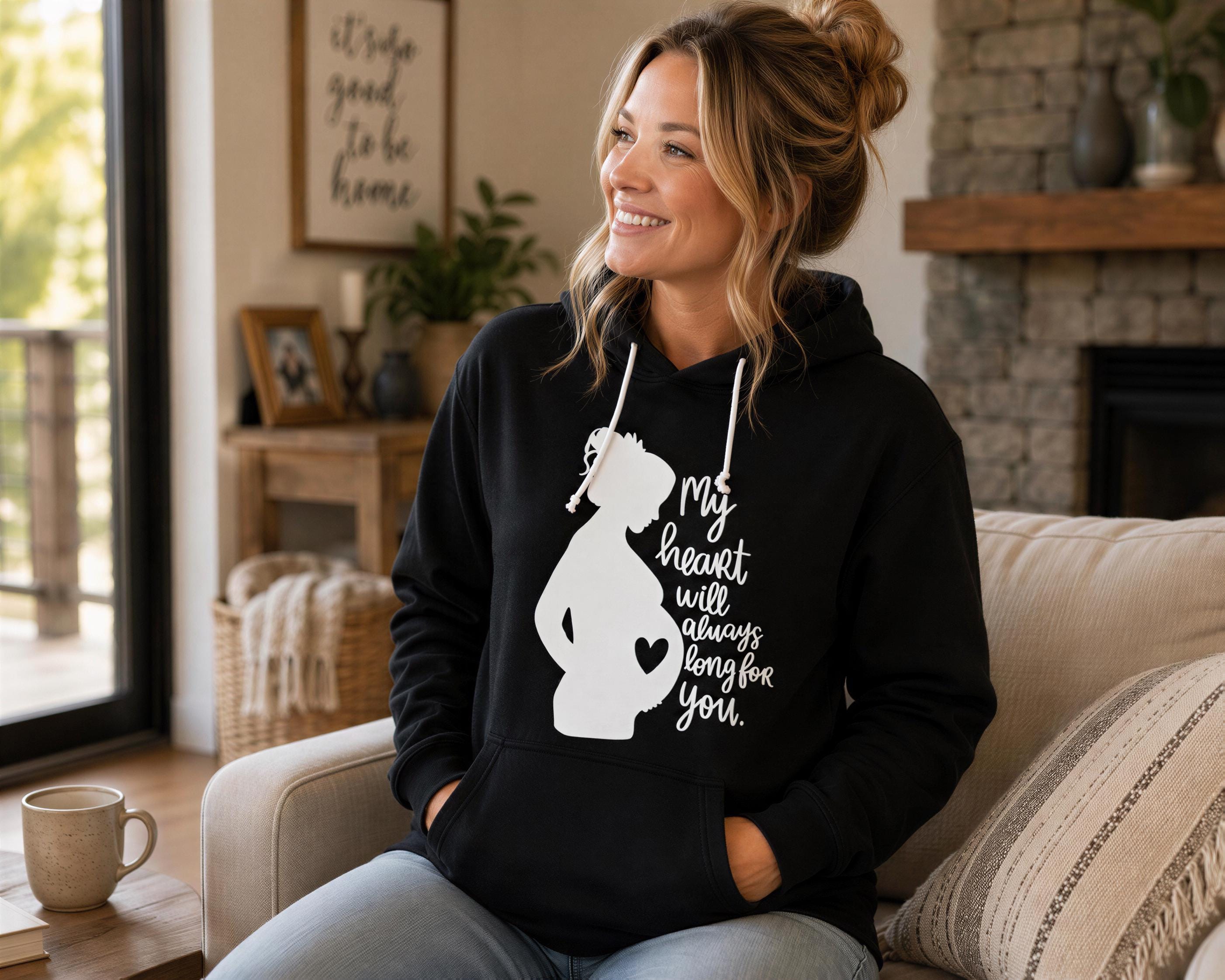 A woman wearing a black hoodie with a white graphic of a pregnant woman and text on it, sitting on a couch in a living room.