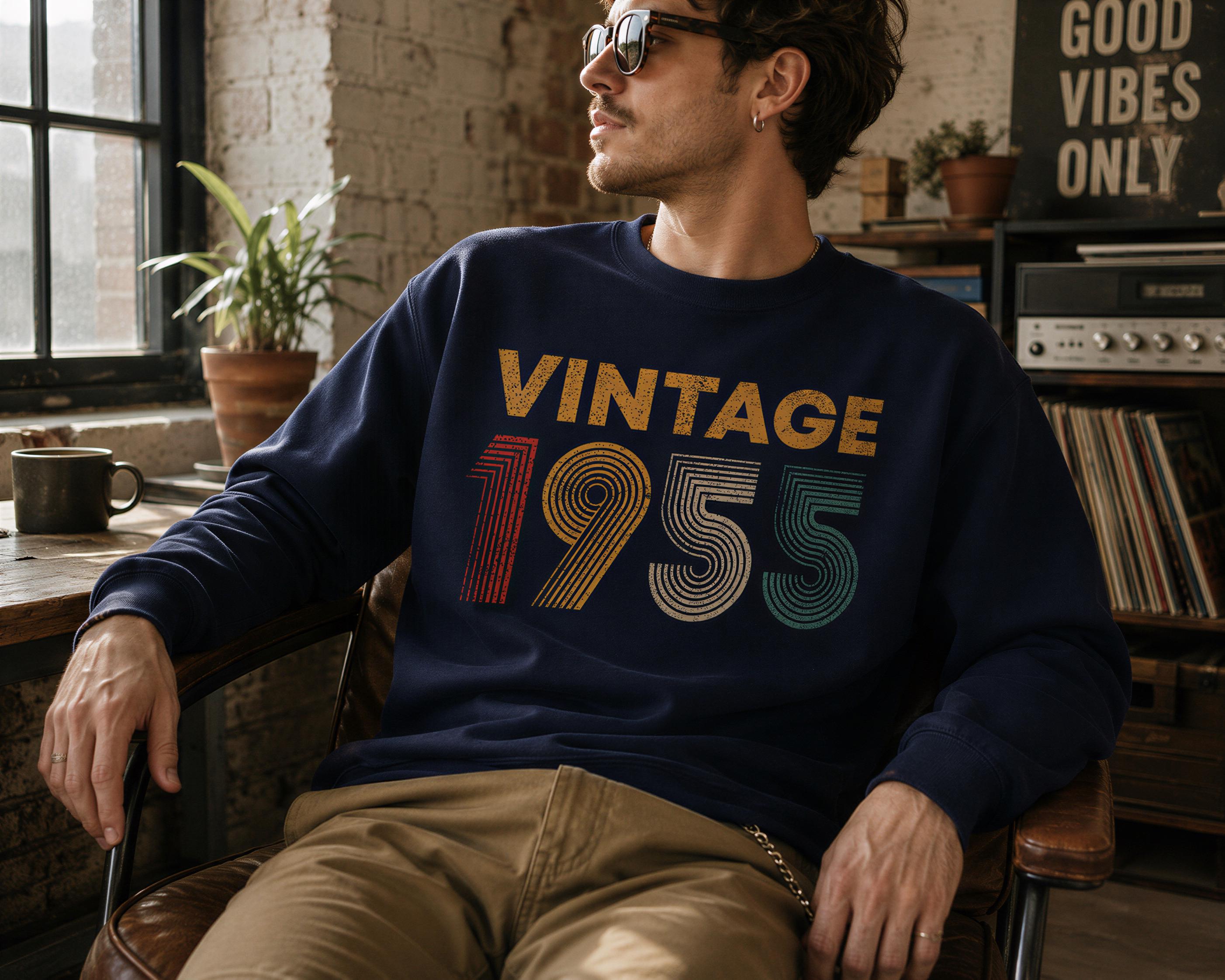 A man wearing a navy blue sweatshirt with the text "VINTAGE 1955" is sitting in a chair in a room with a window, plants, and a record player visible in the background.