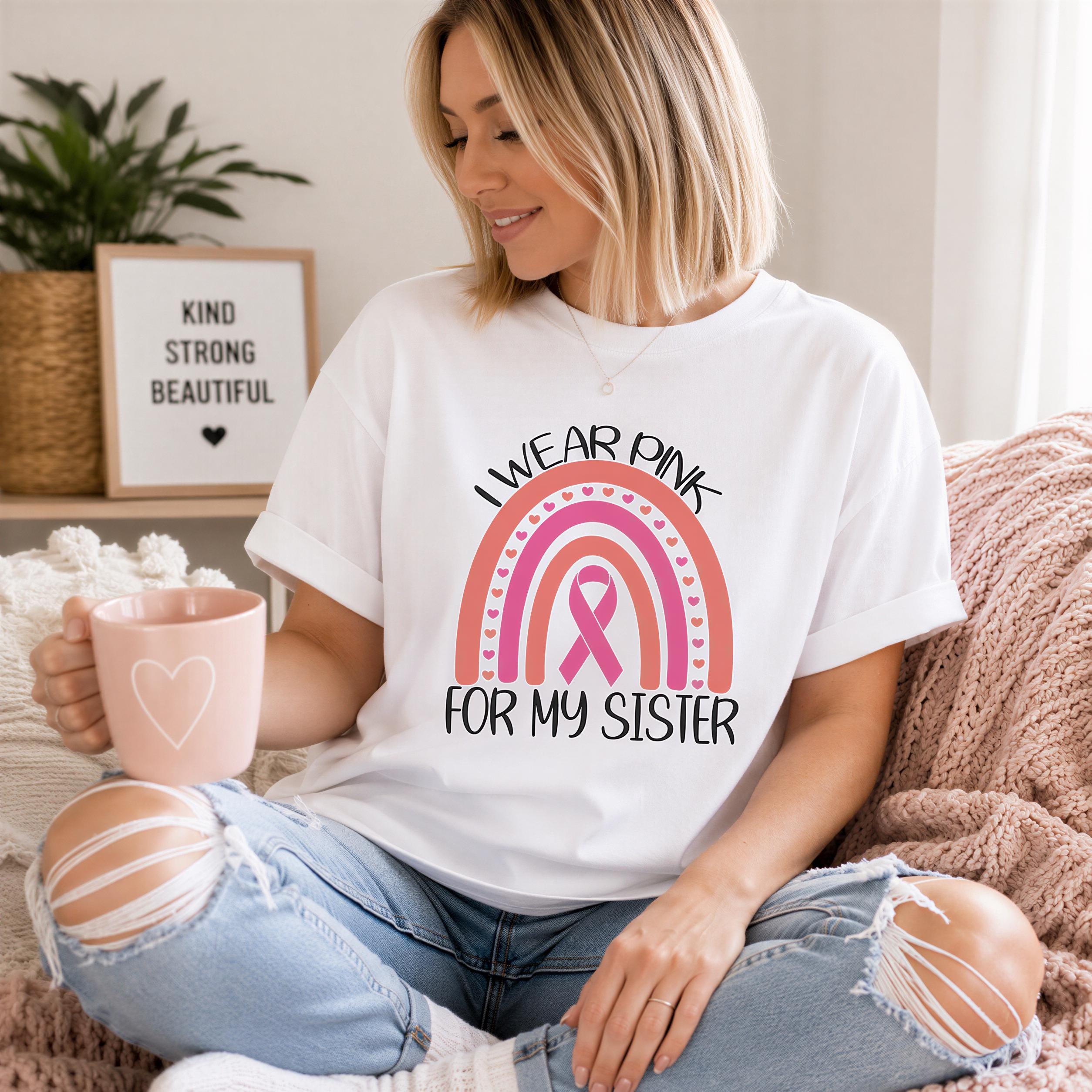 A woman wearing a white t-shirt with a pink ribbon graphic and text, sitting on a couch and holding a pink mug.