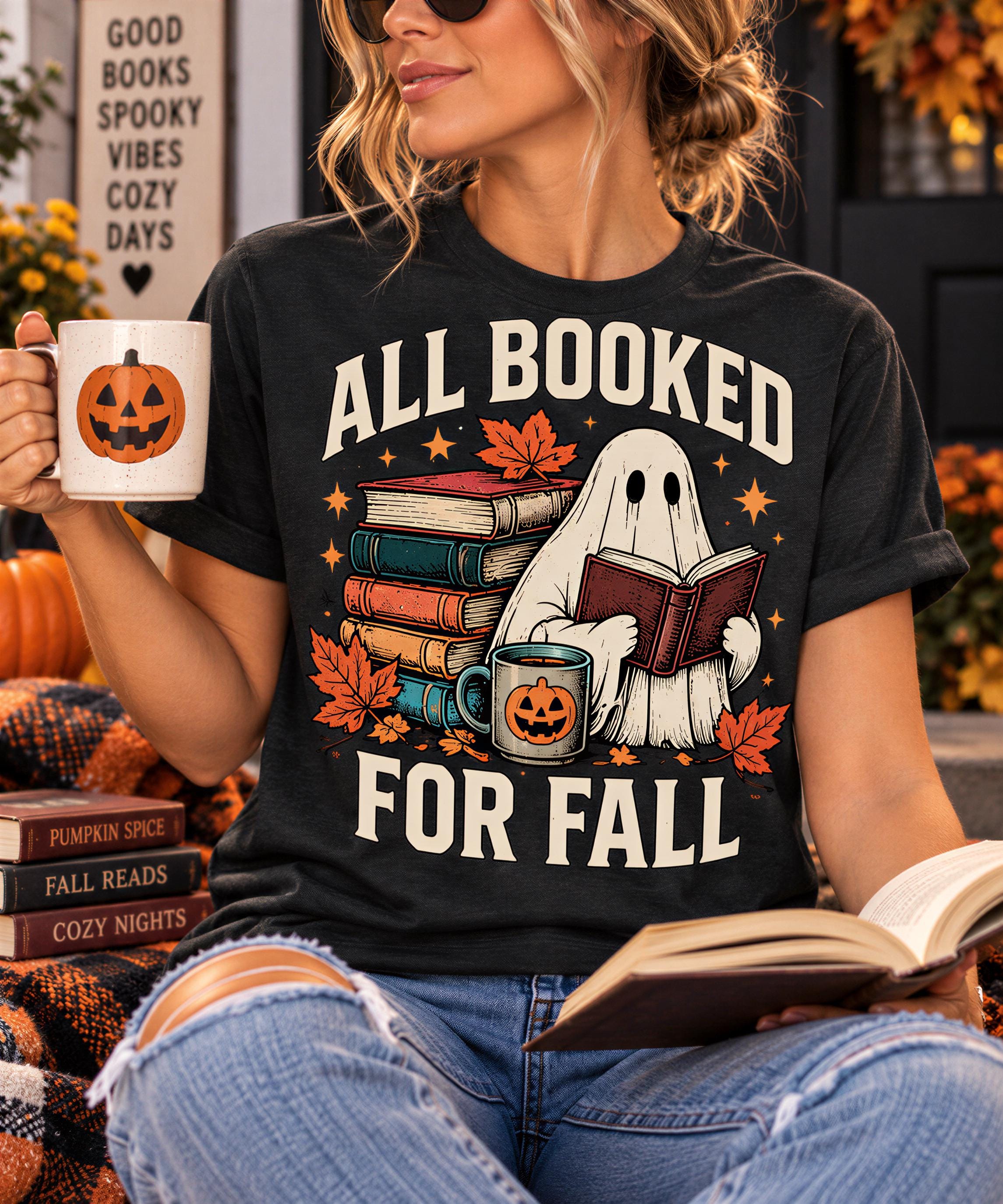 A woman wearing a black t-shirt with a graphic design of a ghost holding a book and a mug, surrounded by autumn leaves and pumpkins, with the text "ALL BOOKED FOR FALL".