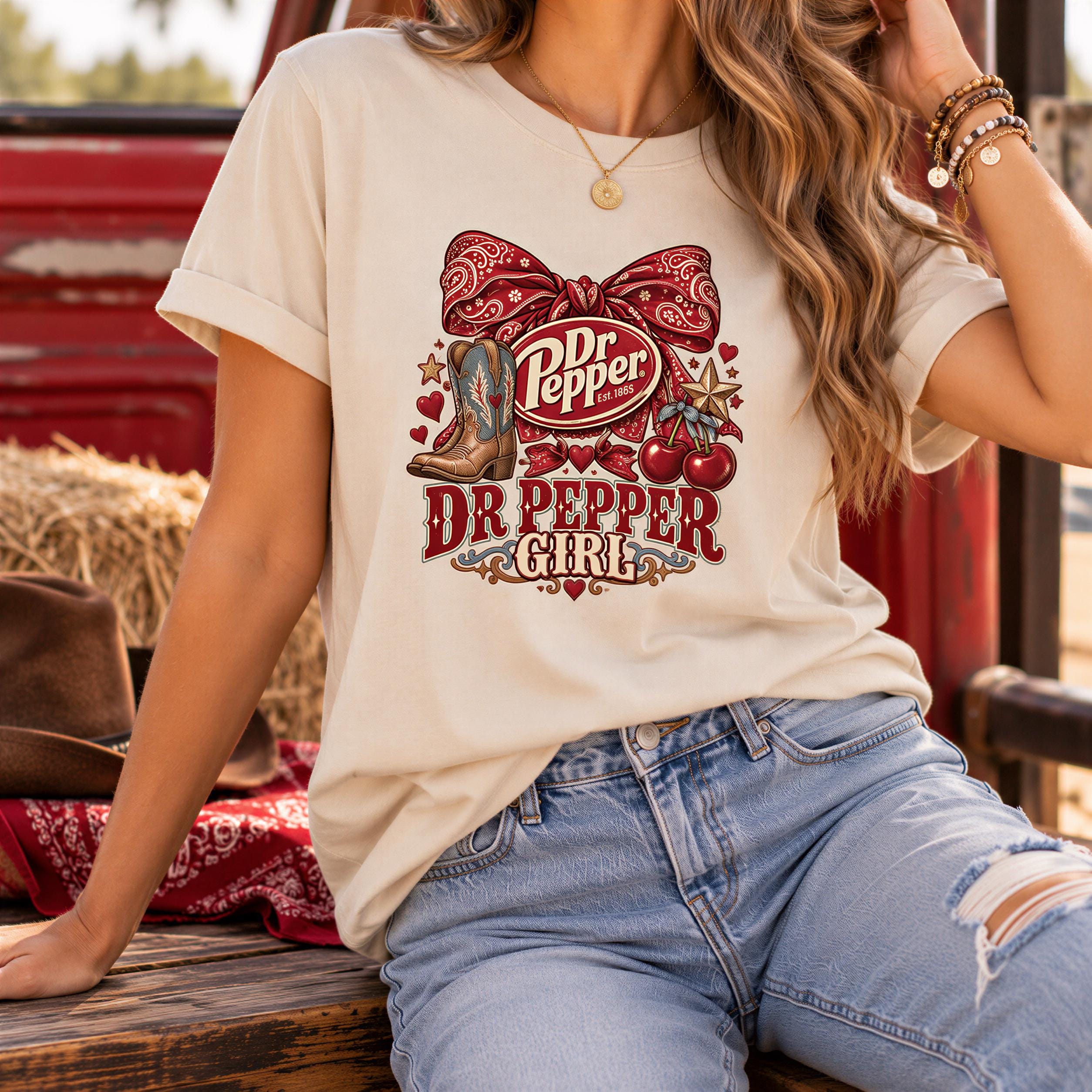 A woman wearing a white t-shirt with a graphic design of Dr Pepper and a bow, paired with blue jeans and a cowboy hat, sitting on a wooden bench.
