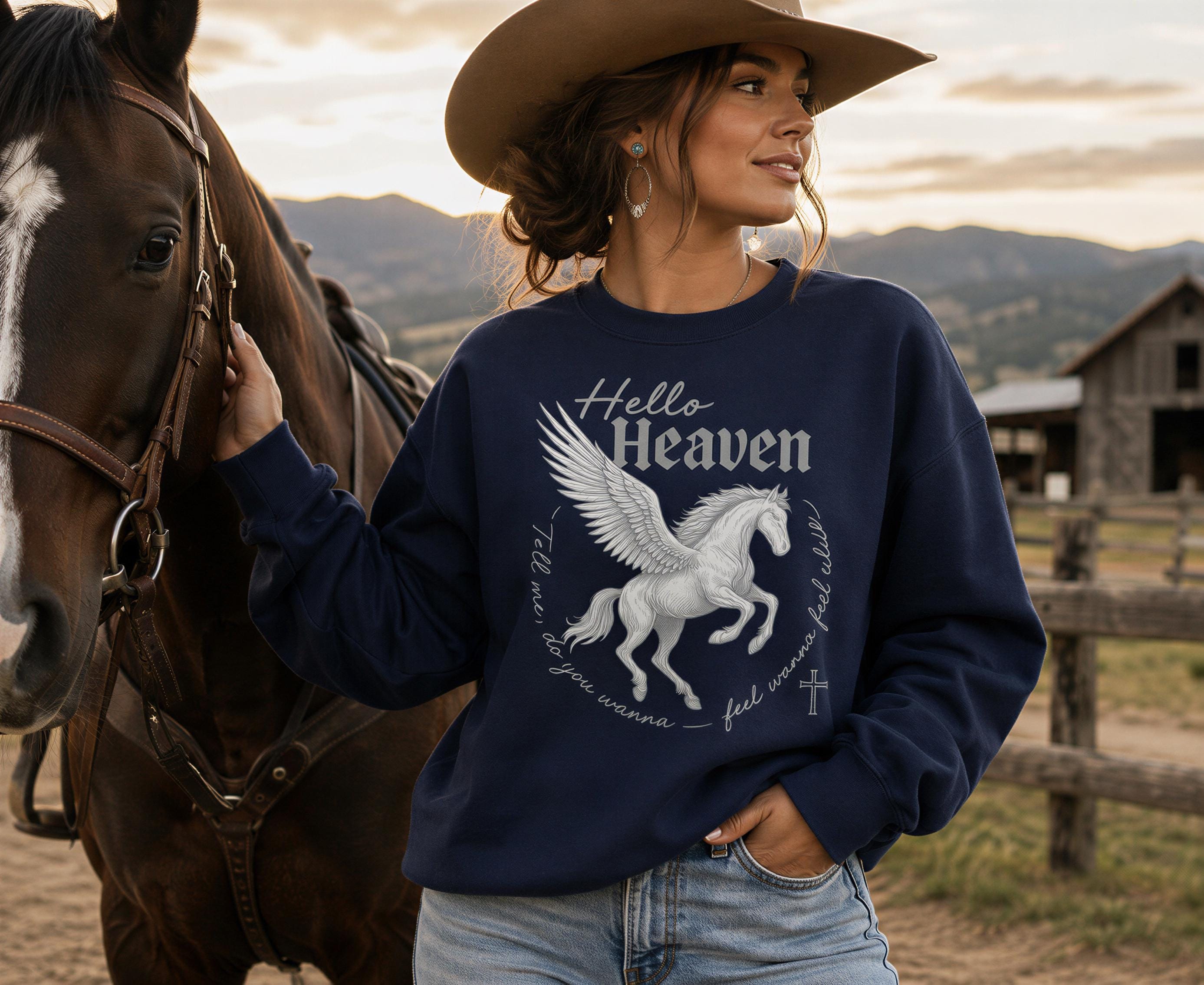 A woman wearing a cowboy hat and a navy blue sweatshirt with a graphic of a winged horse stands next to a brown horse in a rural setting.