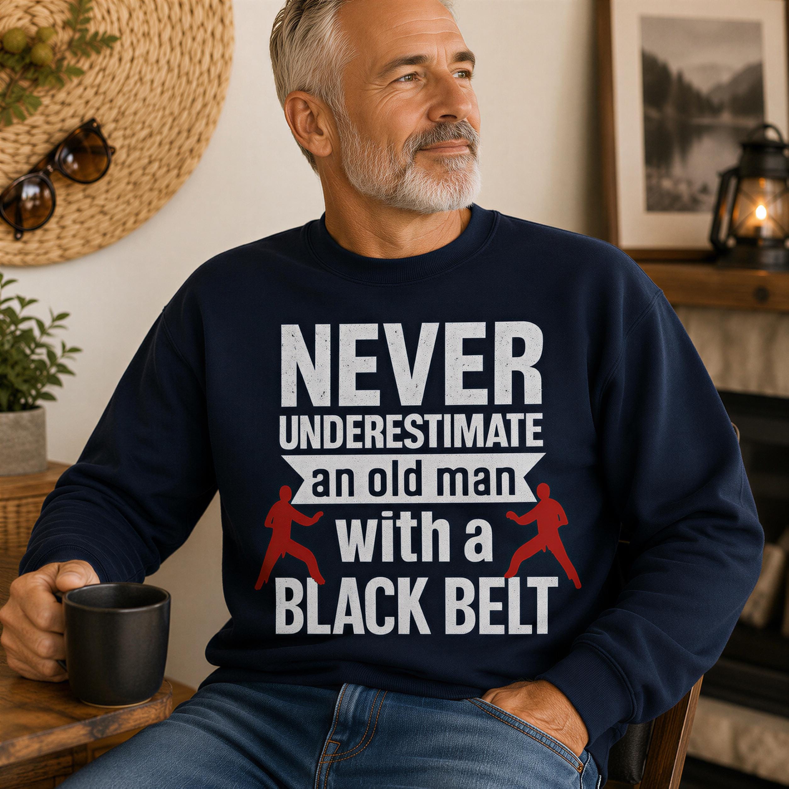 A man wearing a navy blue sweatshirt with the text "NEVER UNDERESTIMATE an old man with a BLACK BELT" is sitting in a room with a potted plant and a framed picture on the wall.