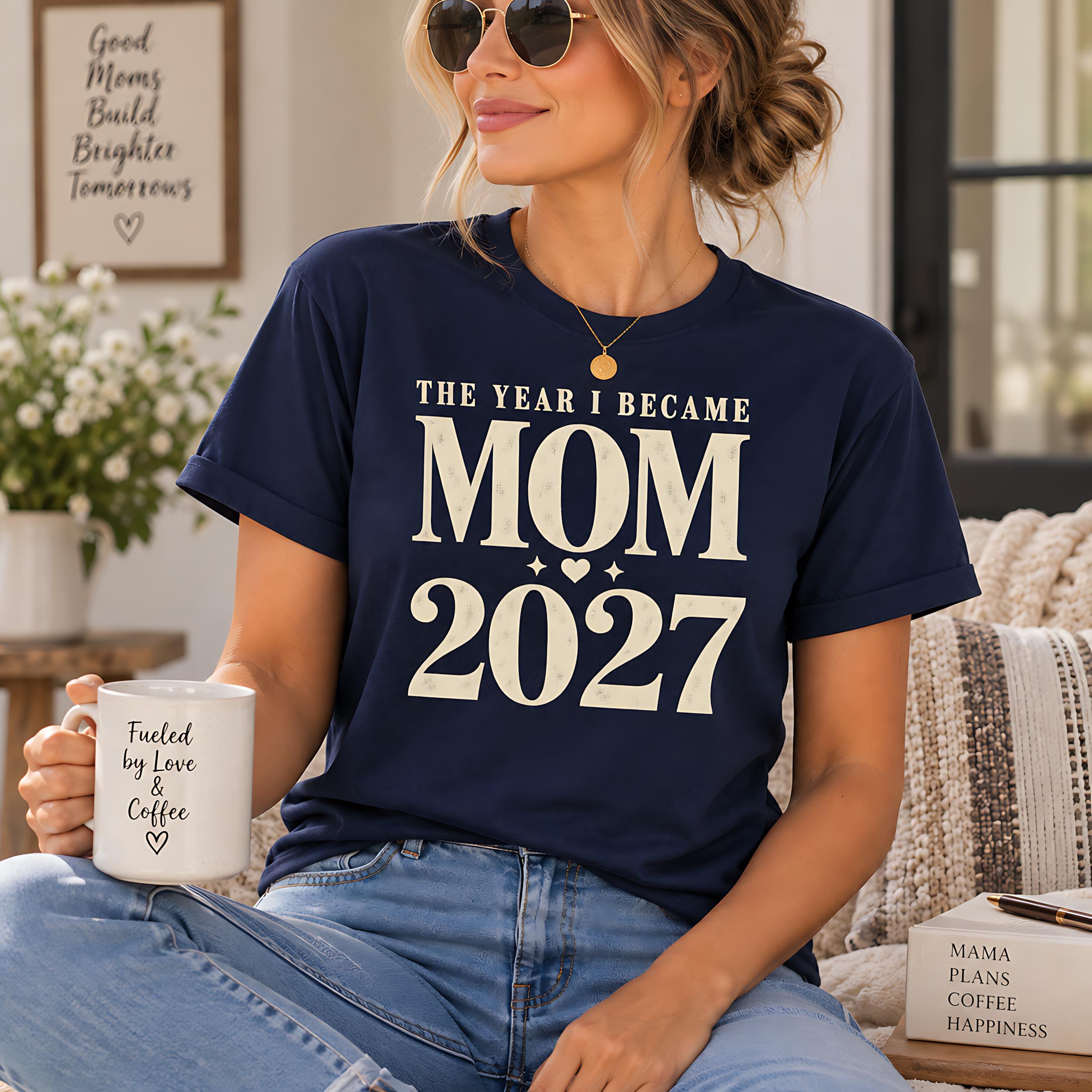 A woman wearing a navy blue t-shirt with the text "The Year I Became Mom 2027" is sitting on a couch, holding a white coffee mug with the text "Founded by love & coffee" printed on it.