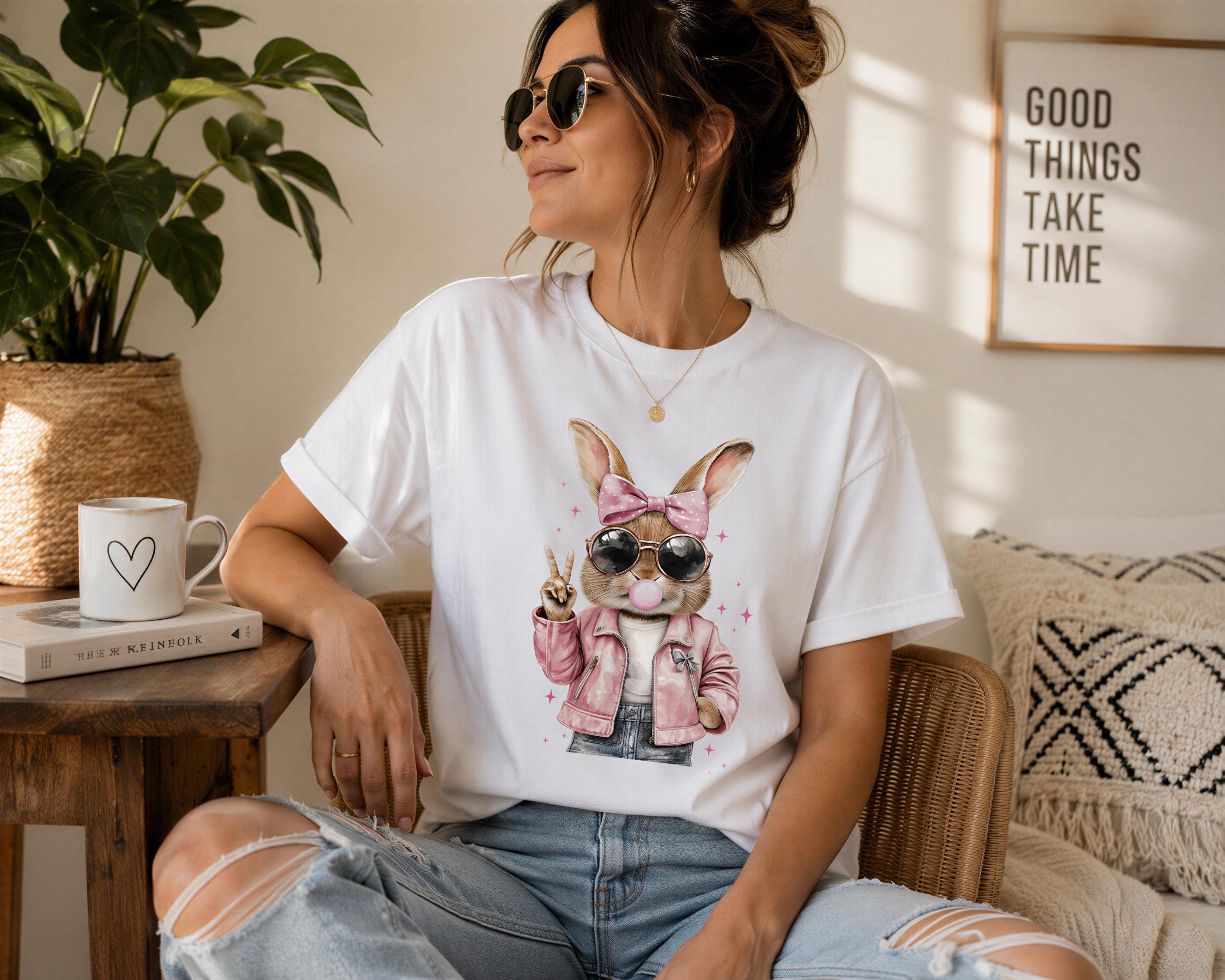 A woman wearing a white t-shirt with a bunny graphic and sunglasses is sitting on a chair in a room with a plant and a framed quote on the wall.
