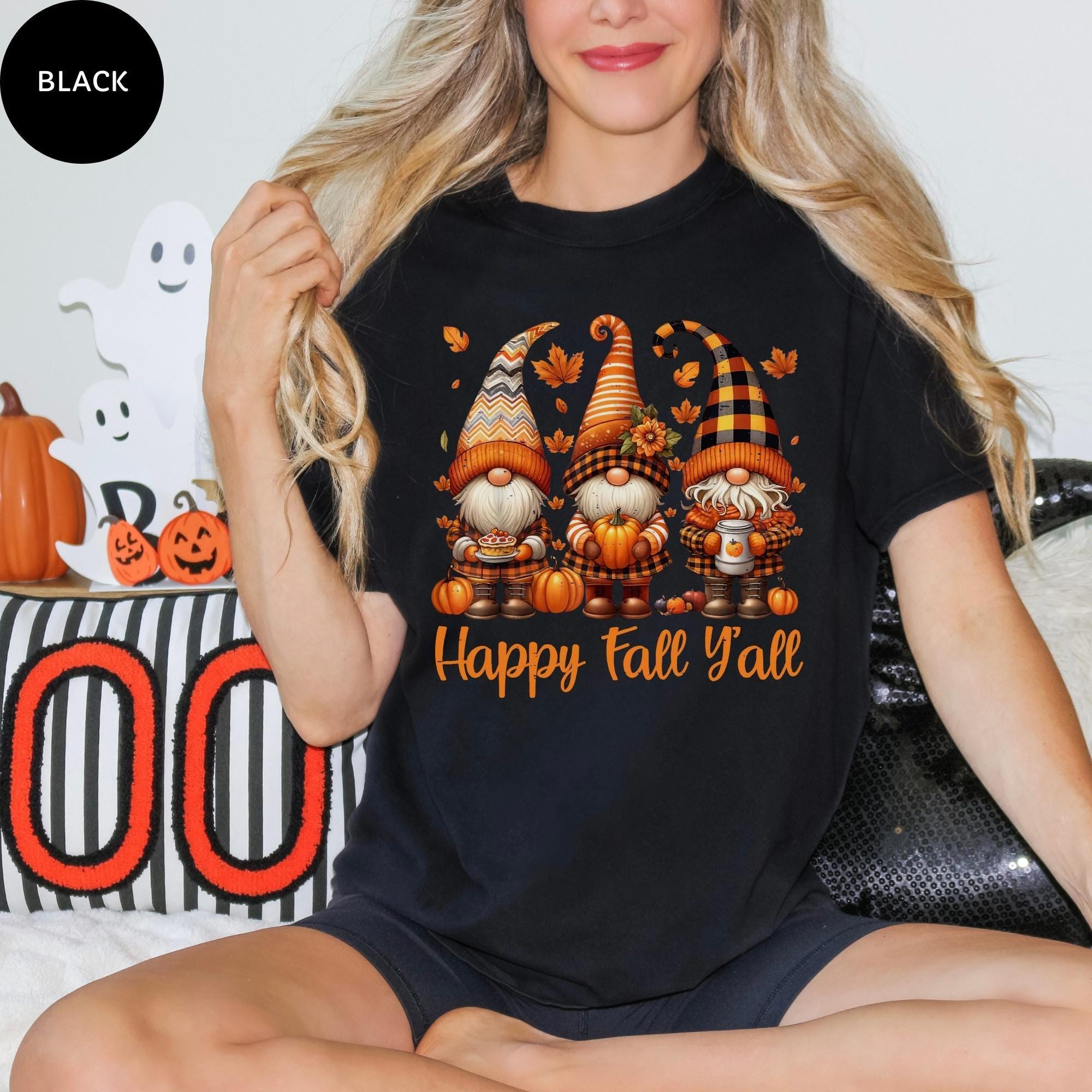 A woman wearing a black t-shirt with a graphic design of three gnomes and the text "Happy Fall Y'all" is sitting on a couch.