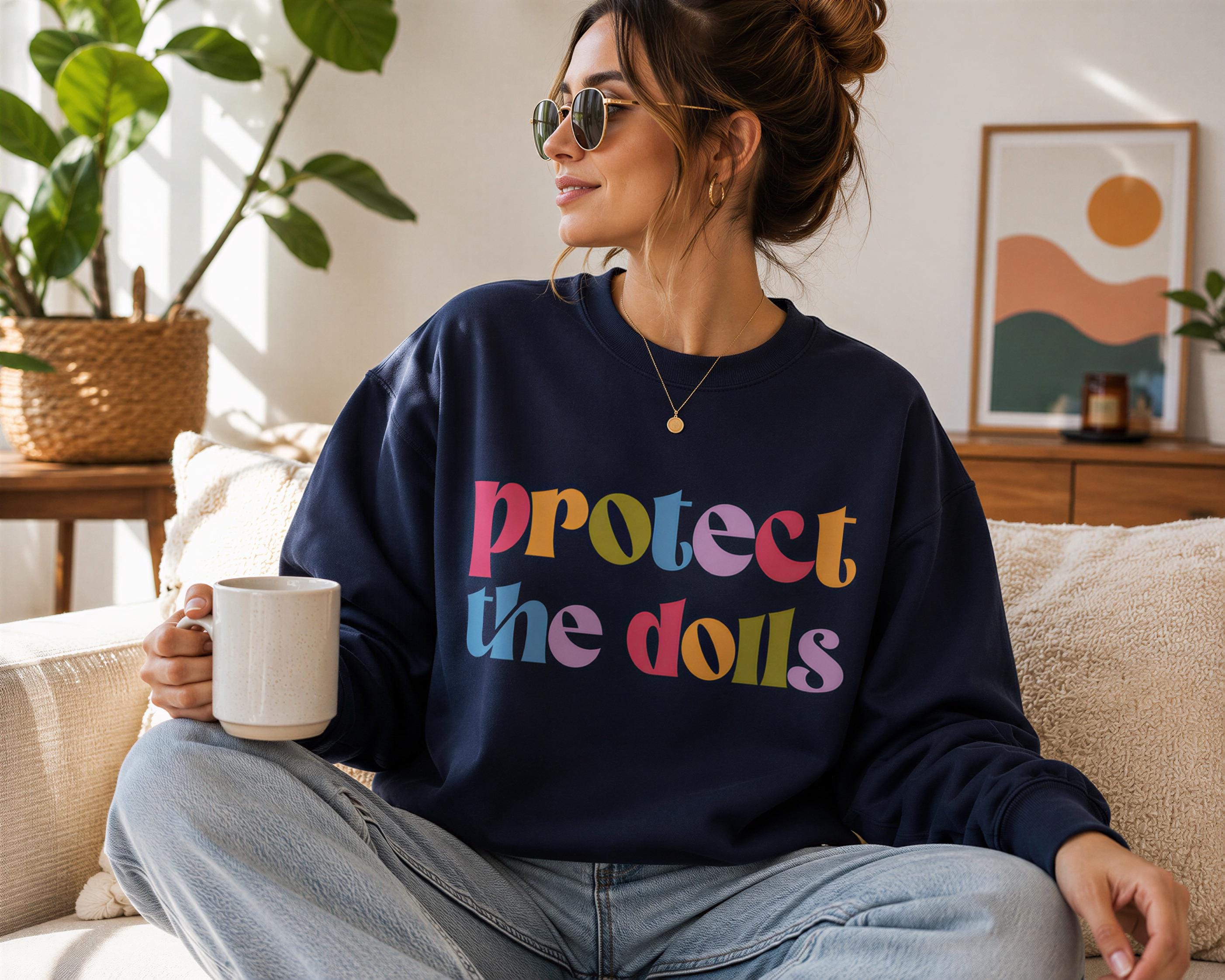 A woman sitting on a couch, wearing a navy blue sweatshirt with the text "Protect the dolls" printed on it, and holding a coffee mug.