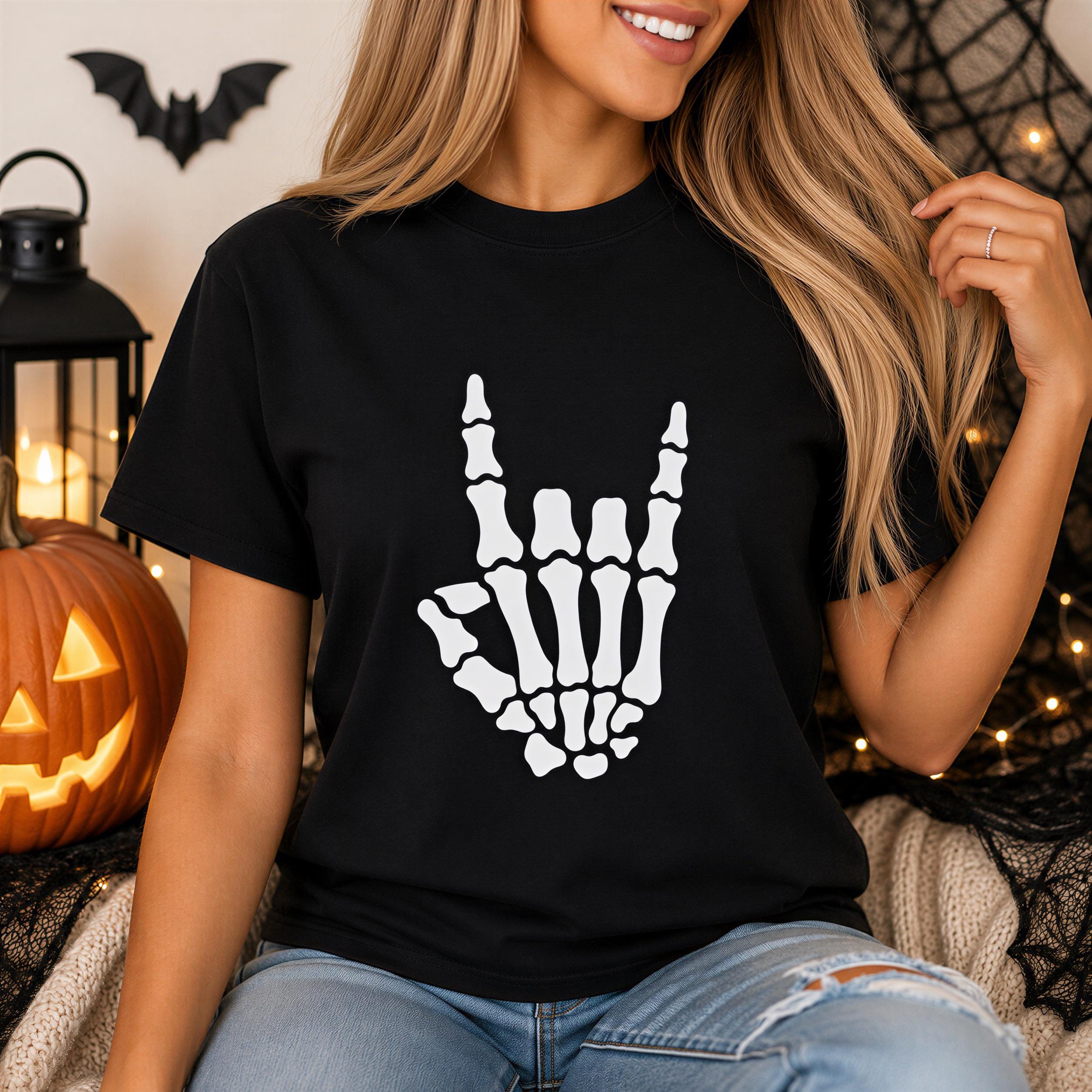 A woman wearing a black t-shirt with a white skeleton hand graphic is sitting in front of a jack-o-lantern and a Halloween decoration.