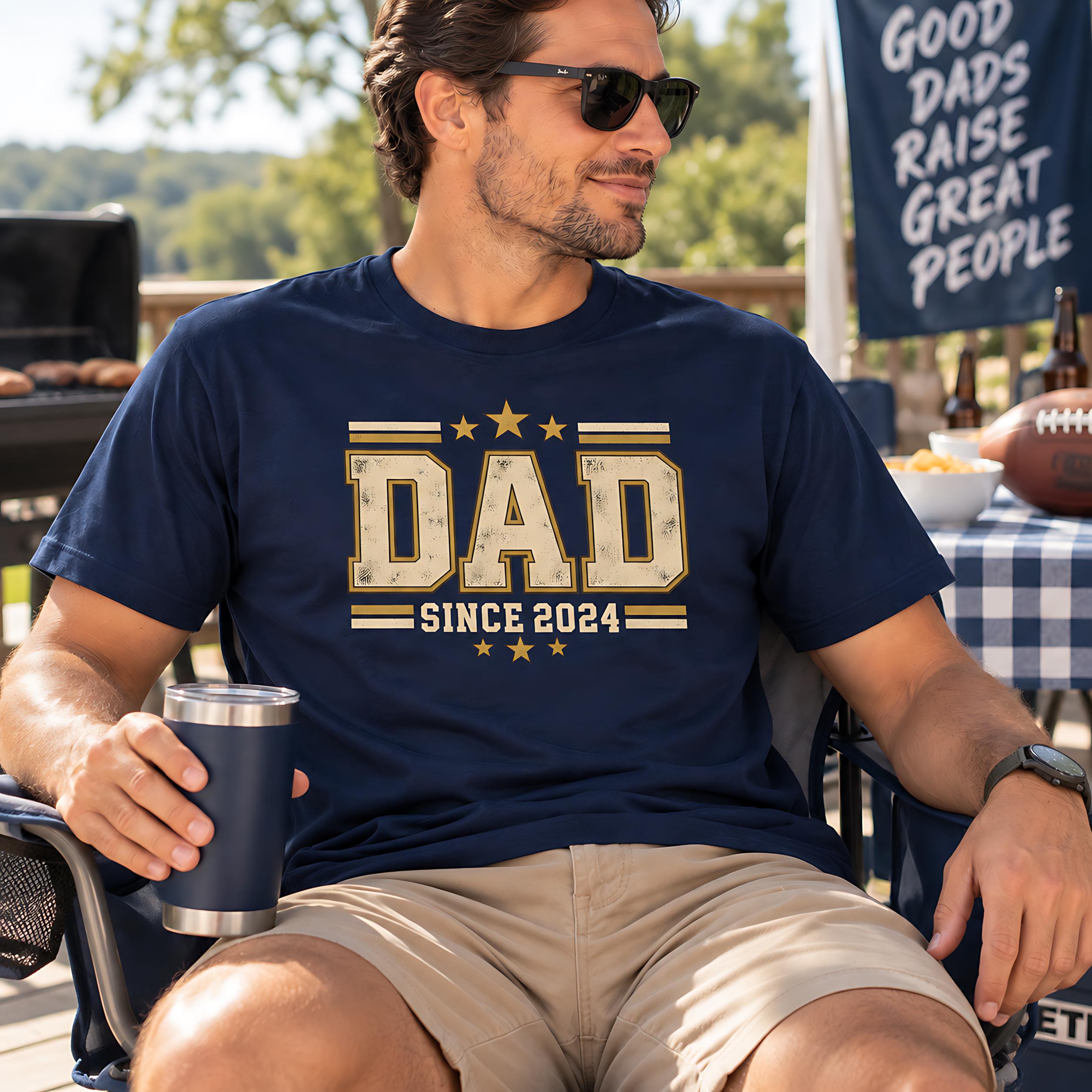 A man wearing a navy blue t-shirt with the text "DAD SINCE 2024" is sitting outdoors, holding a drink and smiling.
