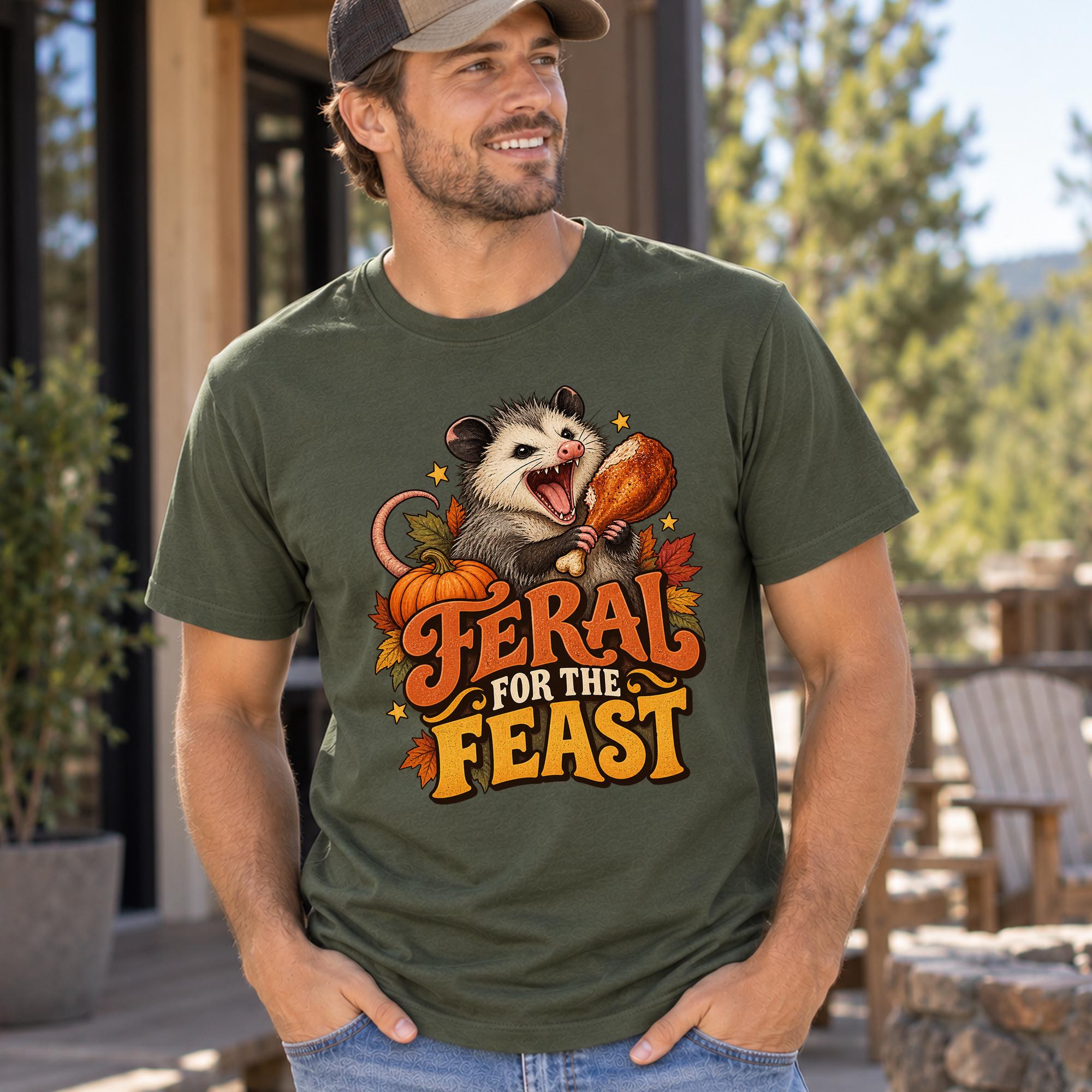 A man wearing a green t-shirt with a graphic design of an opossum holding a turkey and surrounded by autumn leaves, with the text "Feral for the Feast" printed on the shirt.