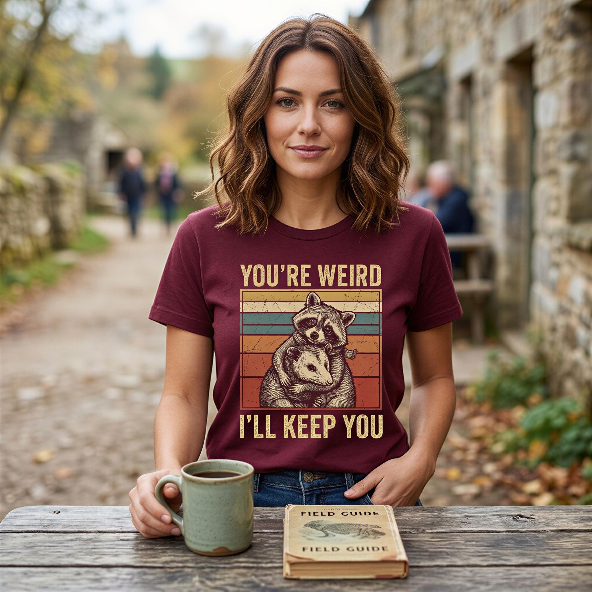 A woman wearing a burgundy t-shirt with a graphic of a raccoon and the text "YOU'RE WEIRD I'LL KEEP YOU" is sitting at a wooden table with a mug of coffee and a book titled "FIELD GUIDE".