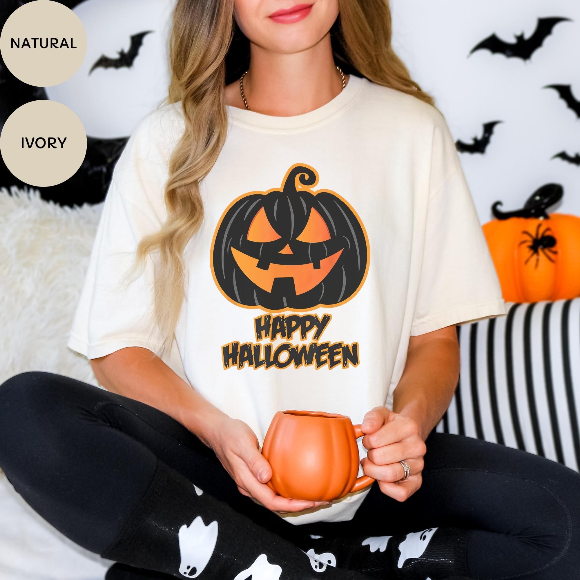A woman wearing a white t-shirt with a jack-o-lantern graphic and the text "Happy Halloween" is sitting on a couch, holding a pumpkin-shaped mug.