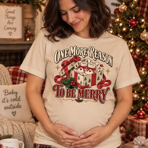 Op de afbeelding: Een beige T-shirt met korte mouwen en een afbeelding van een open geschenkdoos met babyslofjes, omringd door rode linten en hulst. De tekst One More Reason To Be Merry staat in een decoratief lettertype op het T-shirt afgebeeld.