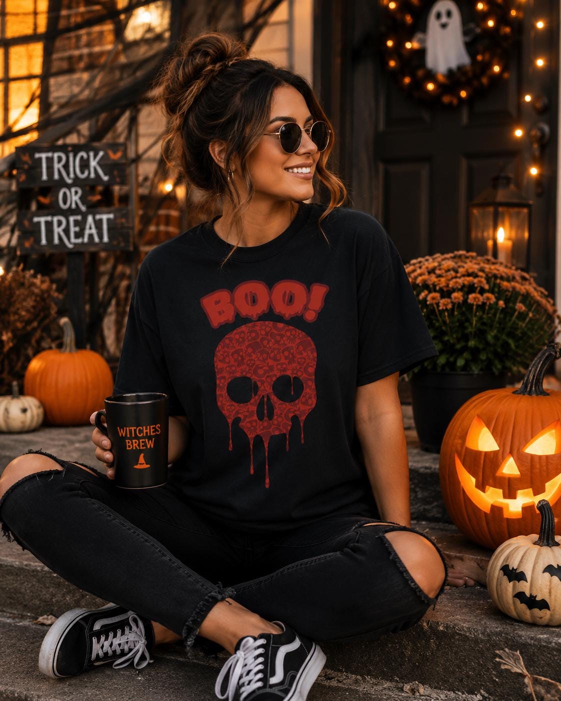 A woman in a black t-shirt with a skull design and the word "BOO!" is sitting on a step, holding a mug with the text "WITCHES BREW" printed on it. Halloween decorations, including pumpkins and a ghost, are visible in the background.