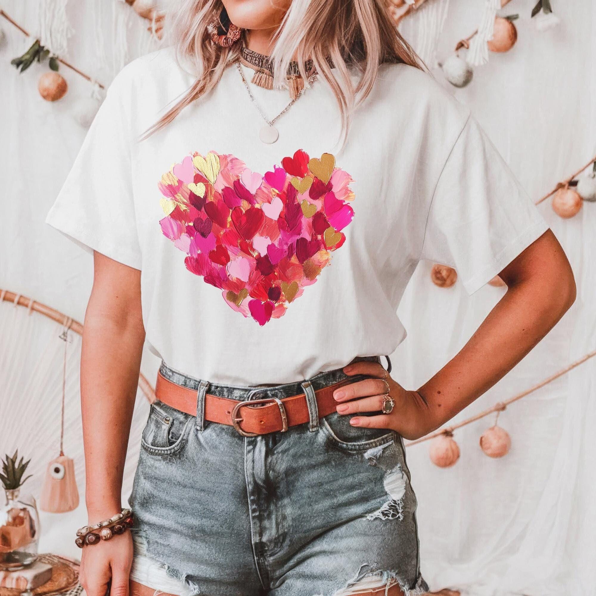 A woman wearing a white t-shirt with a heart design and distressed denim shorts stands in front of a white background with hanging ornaments.
