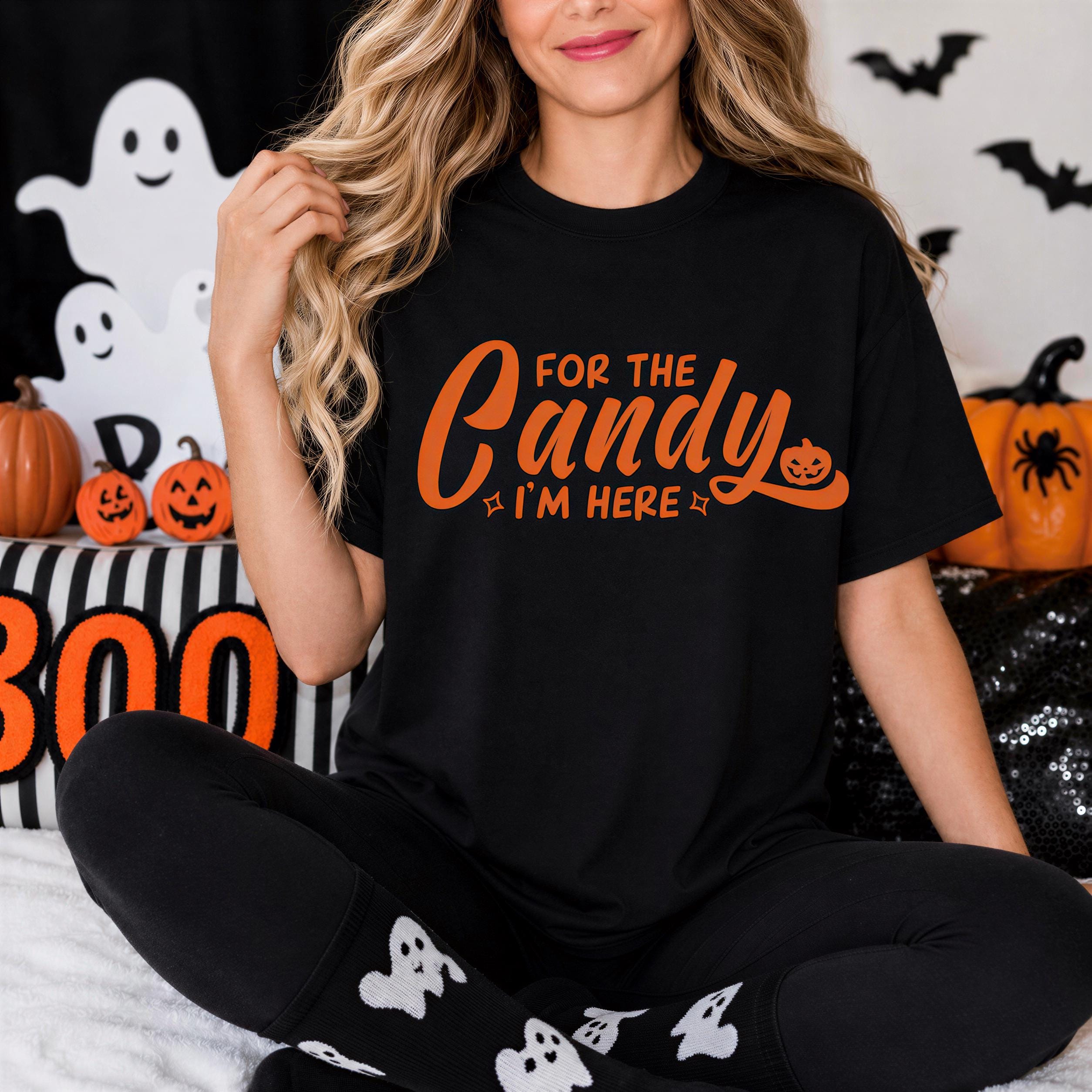 A woman wearing a black t-shirt with the text "FOR THE CANDY I'M HERE" is sitting on a bed surrounded by Halloween decorations, including a ghost, pumpkins, and a "500" sign.