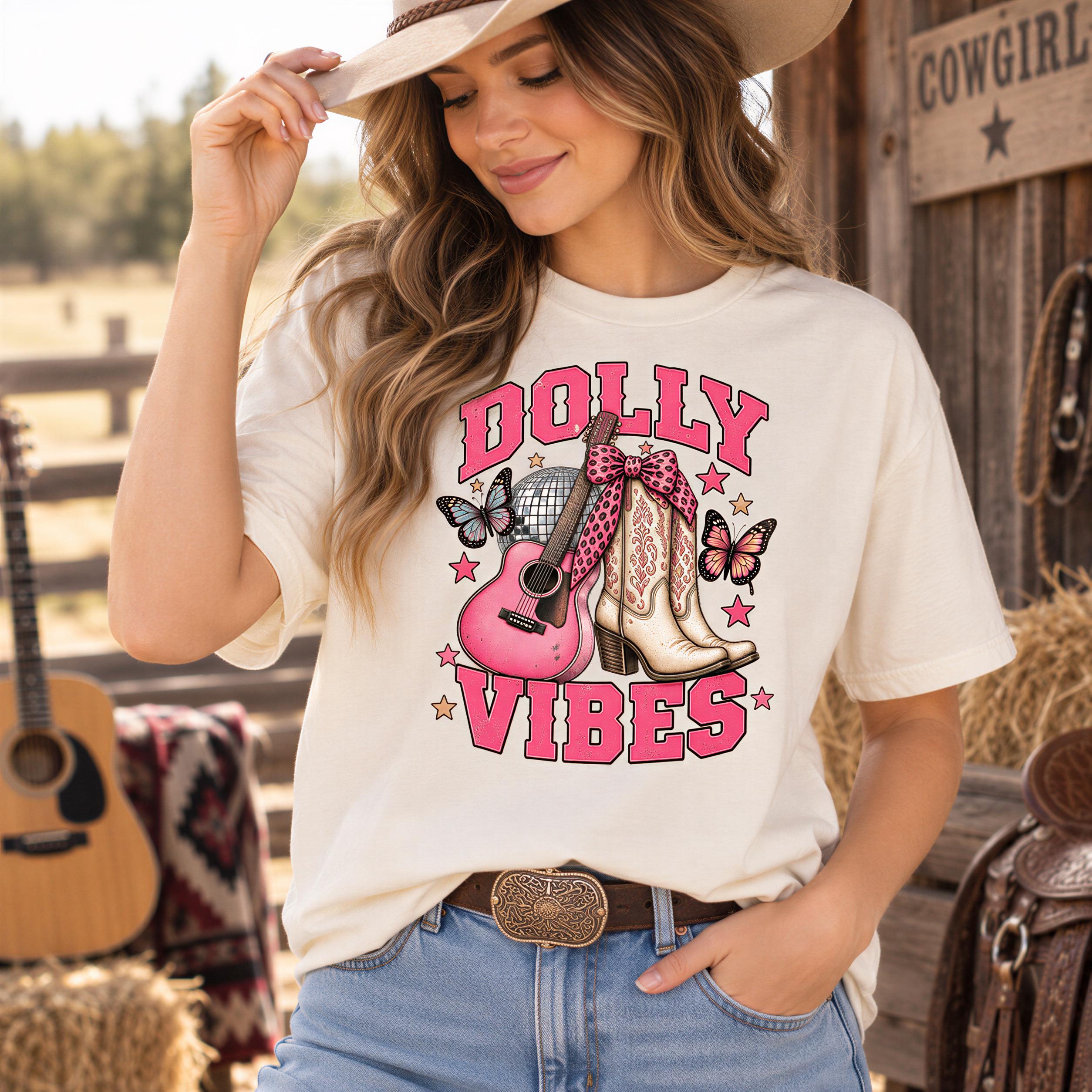 A woman wearing a white t-shirt with a graphic design of a guitar, butterflies, and the text "Dolly Vibes" is standing in front of a wooden structure with a guitar and hay bales.
