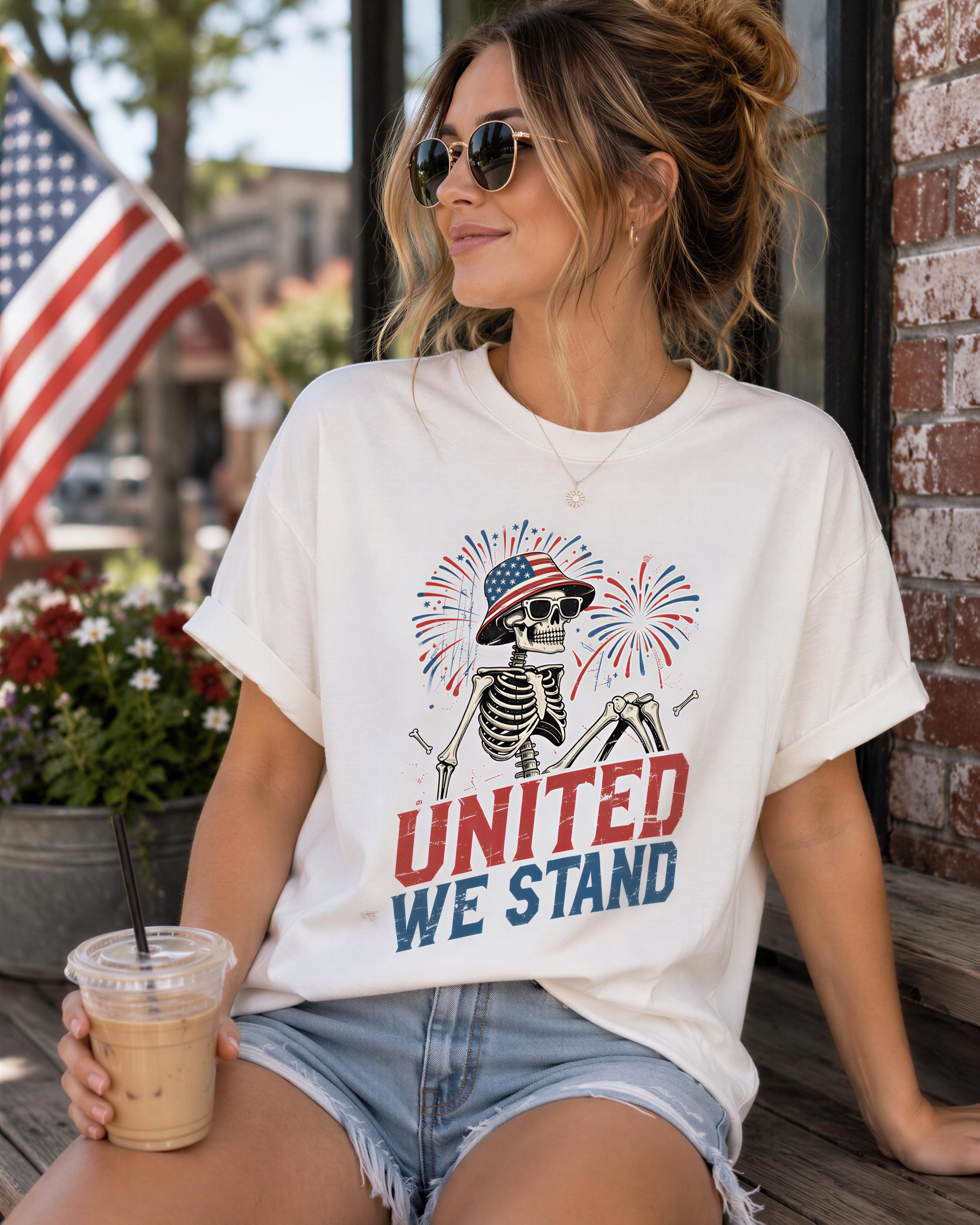 A woman wearing a white t-shirt with a graphic of a skeleton holding a fireworks display and the text "UNITED WE STAND" is sitting on a wooden bench. She is holding a drink in her hand.
