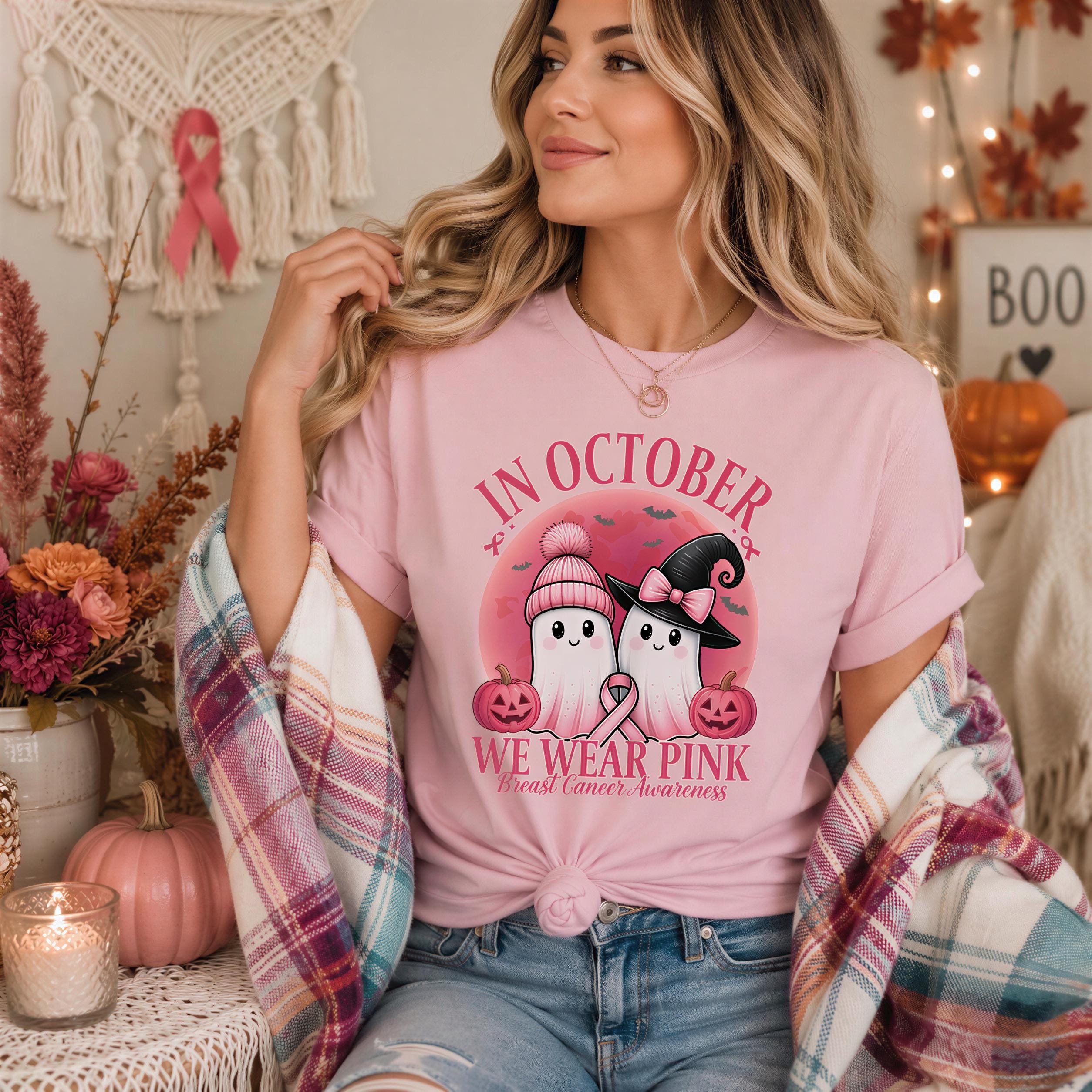 A woman wearing a pink t-shirt with a graphic of two ghostly figures and text, sitting in a room decorated with Halloween-themed items such as pumpkins, candles, and a "BOO" sign.