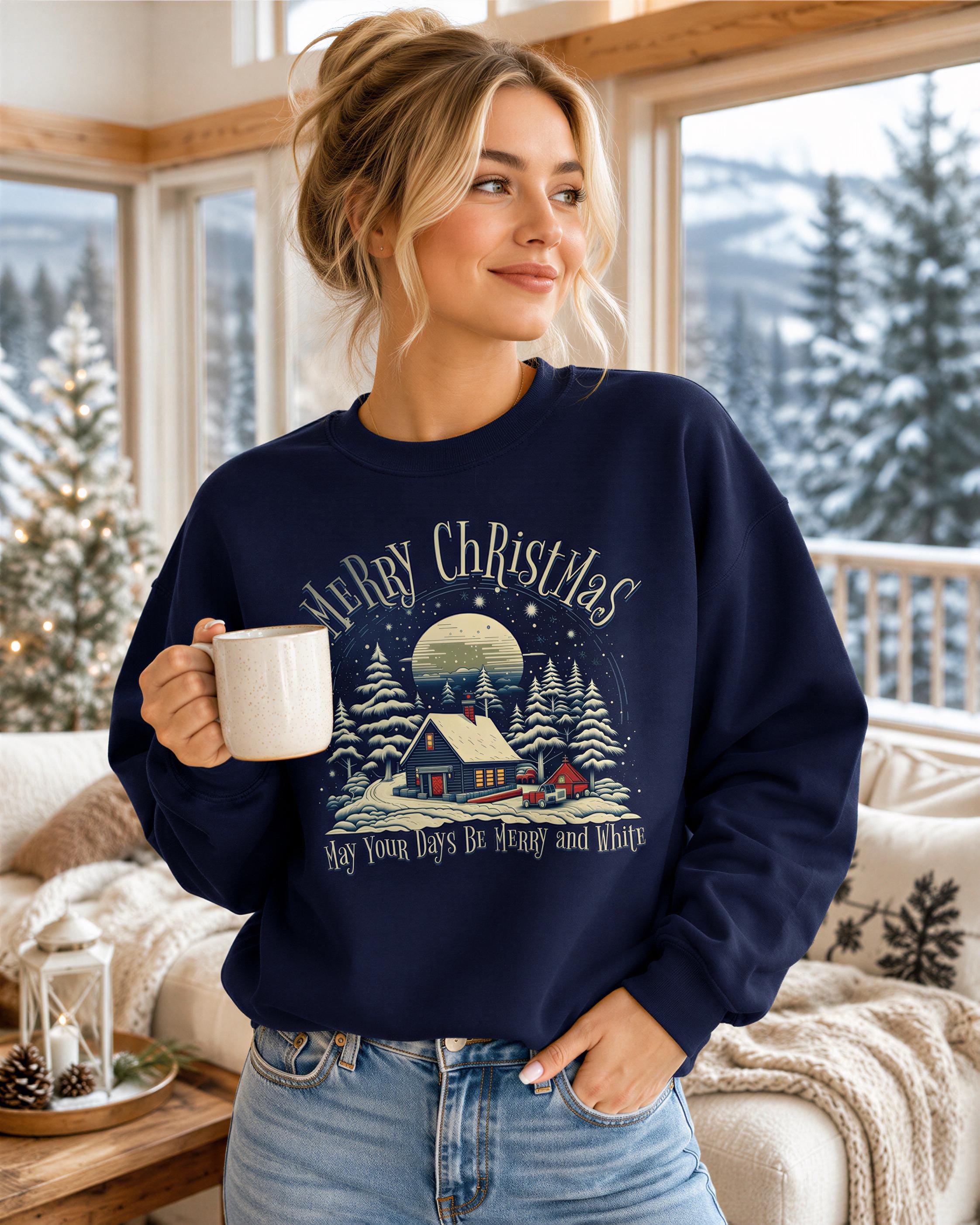 A woman wearing a navy blue sweatshirt with a Christmas-themed design holding a mug, standing in a cozy indoor setting with a window and pine trees visible outside.