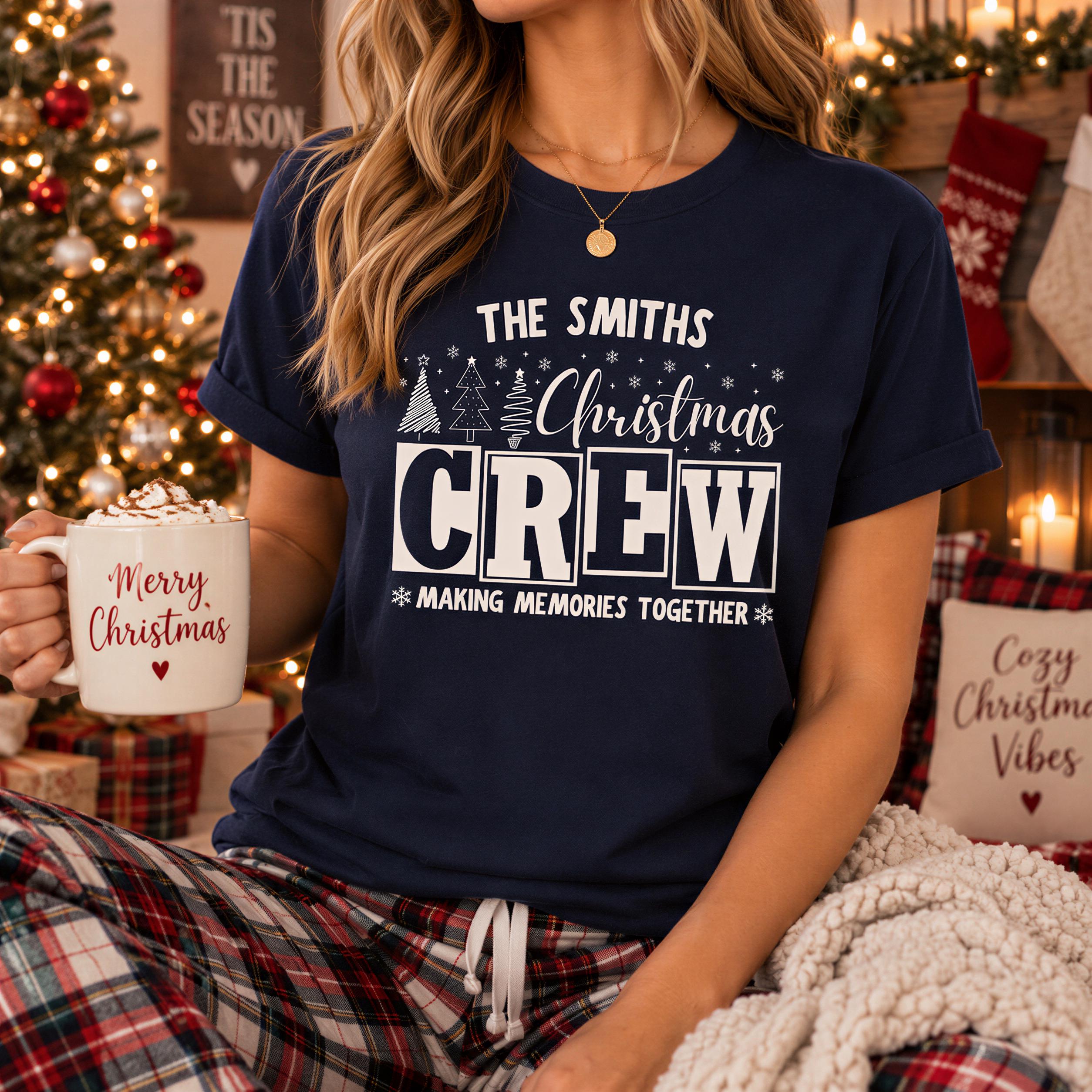 A woman wearing a navy blue t-shirt with a Christmas-themed design, holding a mug with the text "Merry Christmas Crew" and "Making memories together" printed on it. She is sitting in front of a Christmas tree and other holiday decorations.