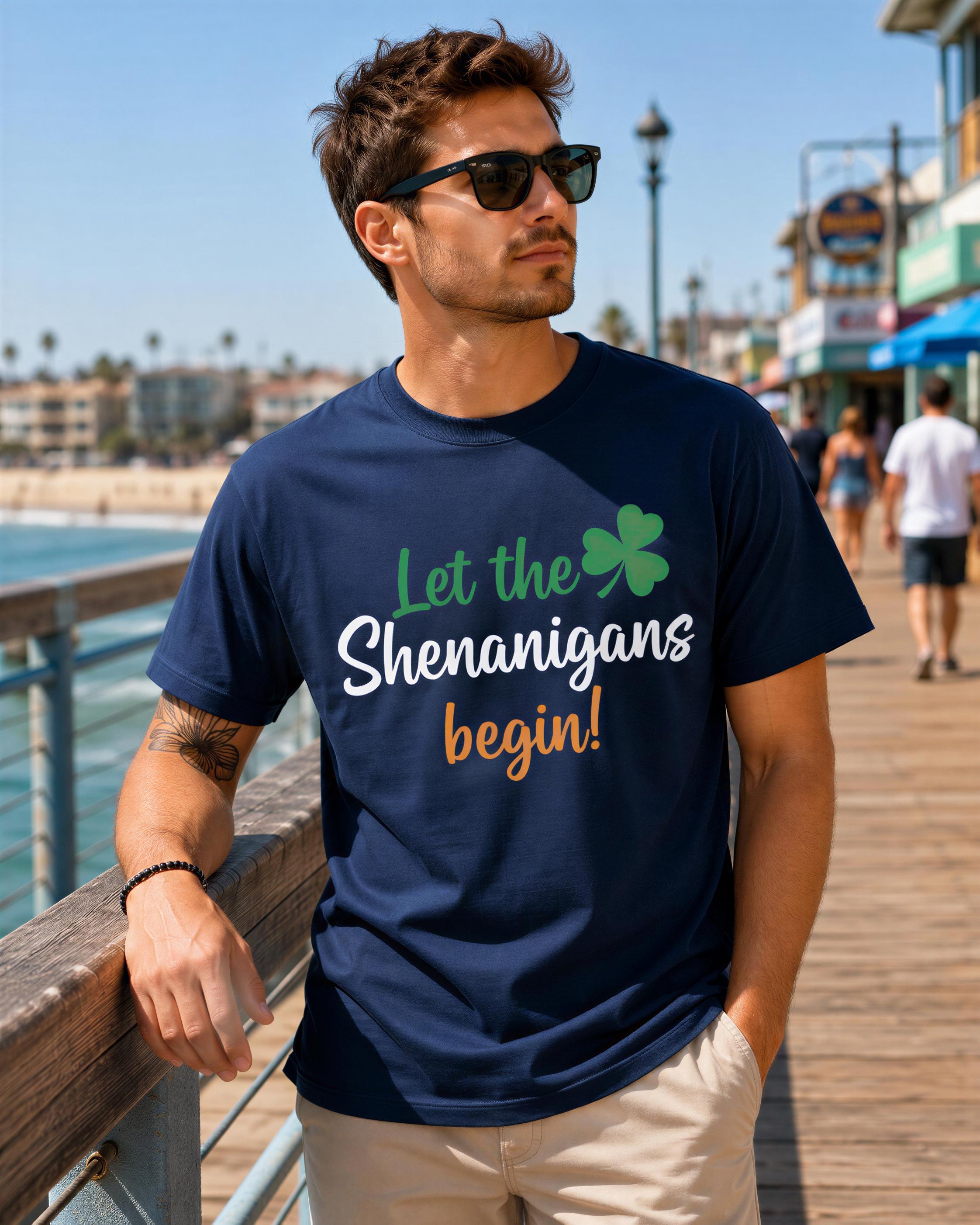 A man wearing a navy blue t-shirt with the text "Let the Shenanigans begin!" is standing on a pier overlooking the ocean.