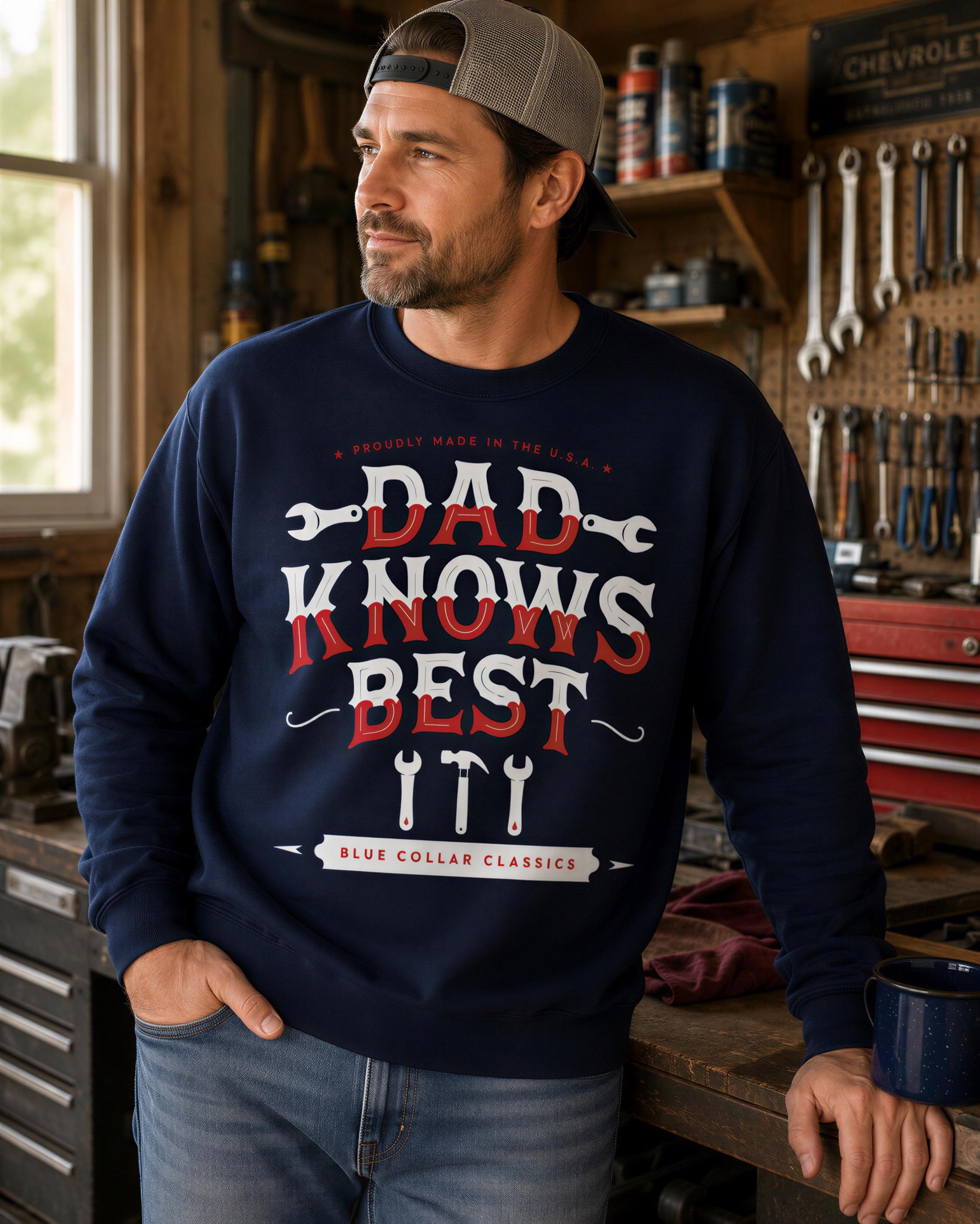 A man wearing a navy blue sweatshirt with a humorous text printed on it, standing in a workshop with various tools and equipment visible in the background.