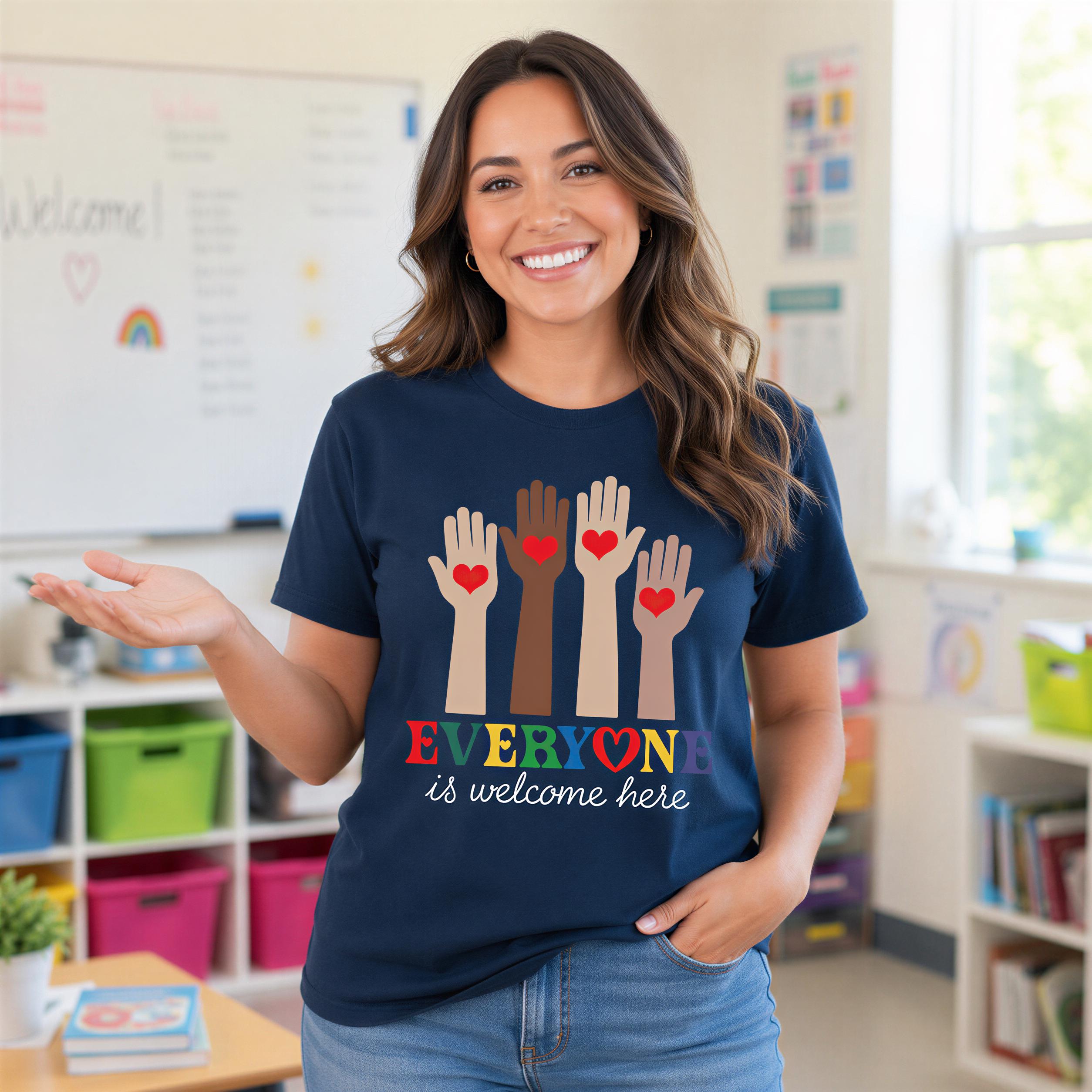 A smiling woman wearing a blue t-shirt with a graphic design of hands holding hearts and the text "EVERYONE is welcome here" in a classroom setting.
