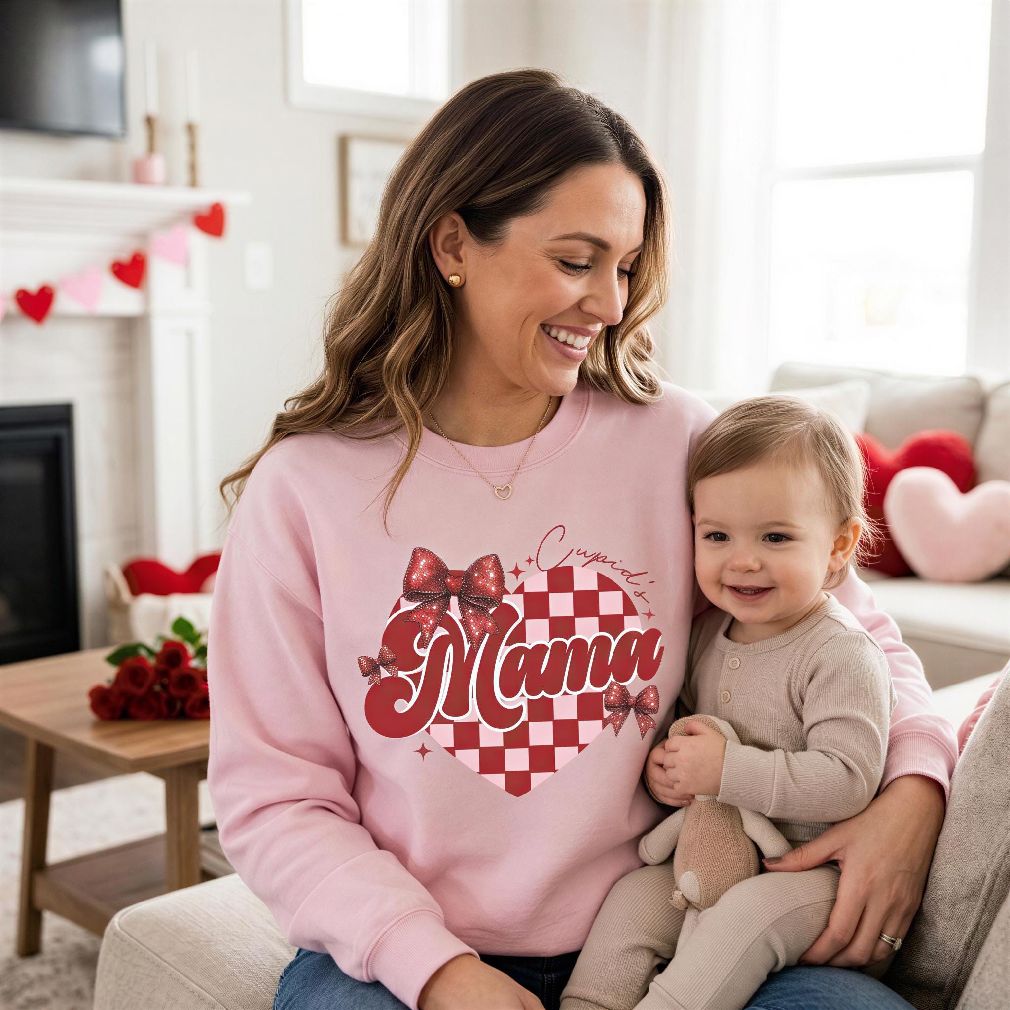 A woman in a pink sweatshirt with a red and white design is holding a smiling baby in a cozy living room with a fireplace, flowers, and a red heart-shaped pillow.