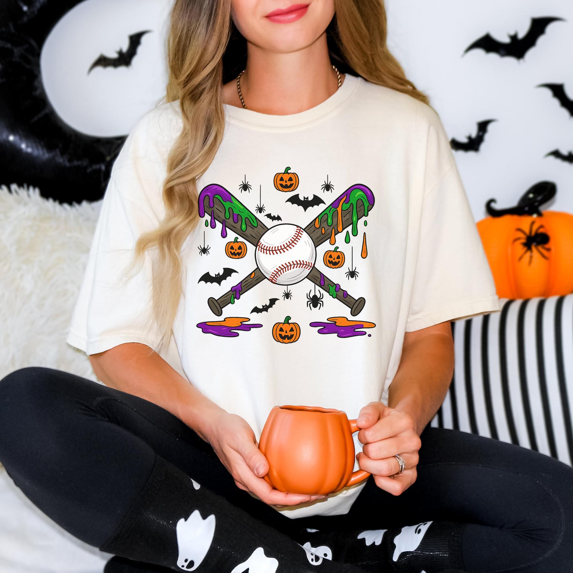 A woman wearing a white t-shirt with a Halloween-themed graphic of baseball bats and a baseball, holding a pumpkin-shaped mug.