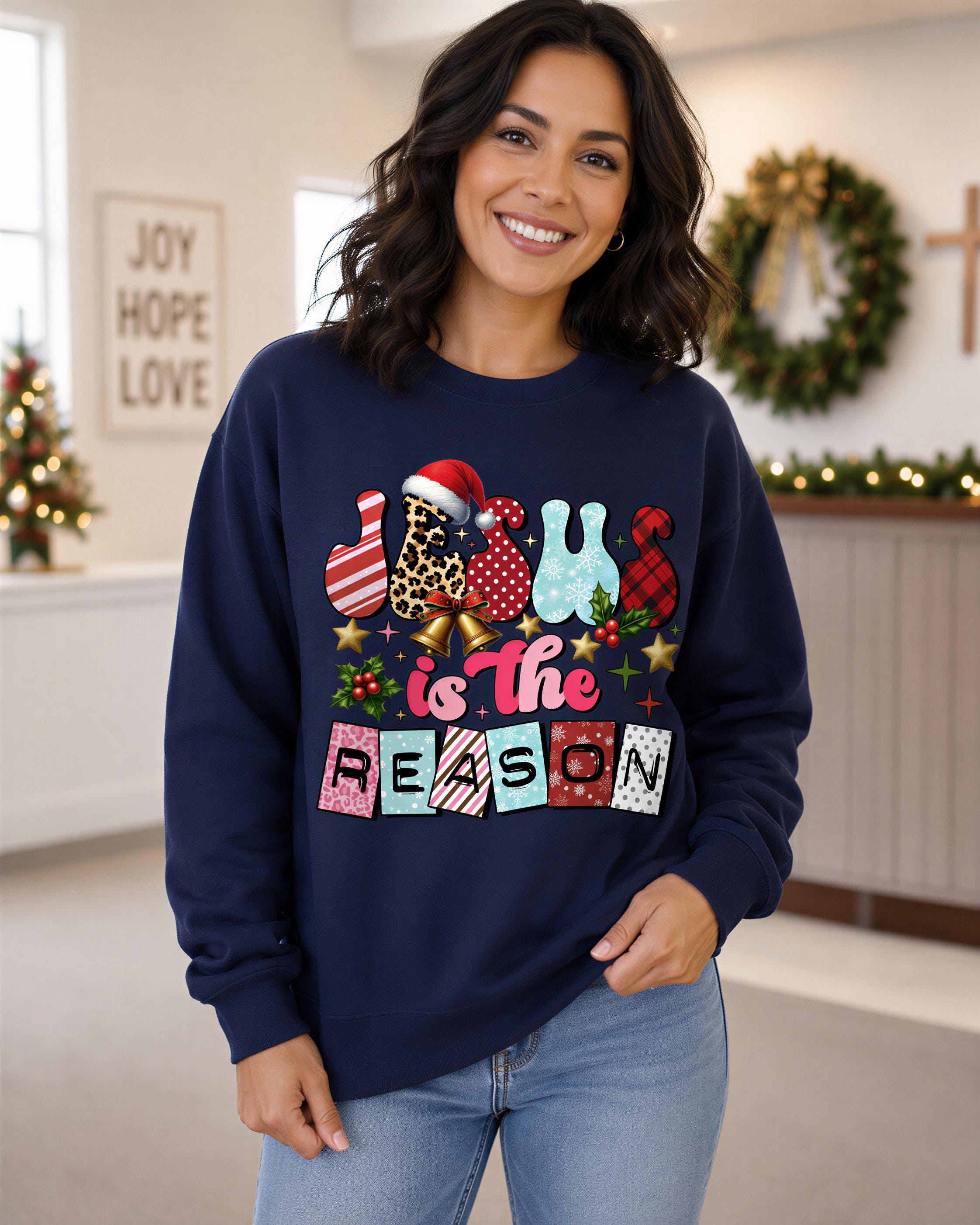 A woman wearing a navy blue sweatshirt with a colorful graphic design that says "JESUS IS THE REASON" and includes various Christmas-themed elements such as a Santa hat, snowflakes, and a wreath.