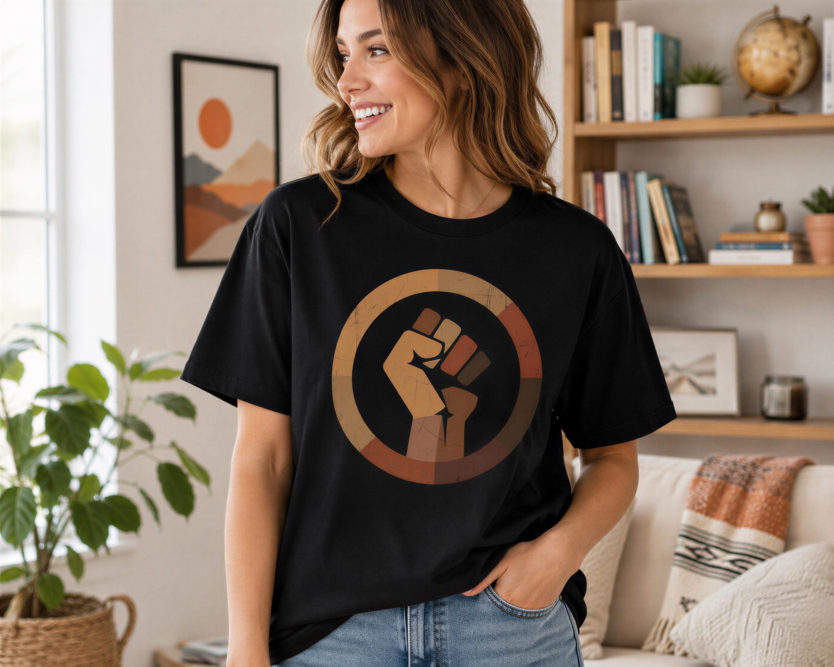 A woman wearing a black t-shirt with a graphic of a fist is standing in a room with a bookshelf and a plant in the background.
